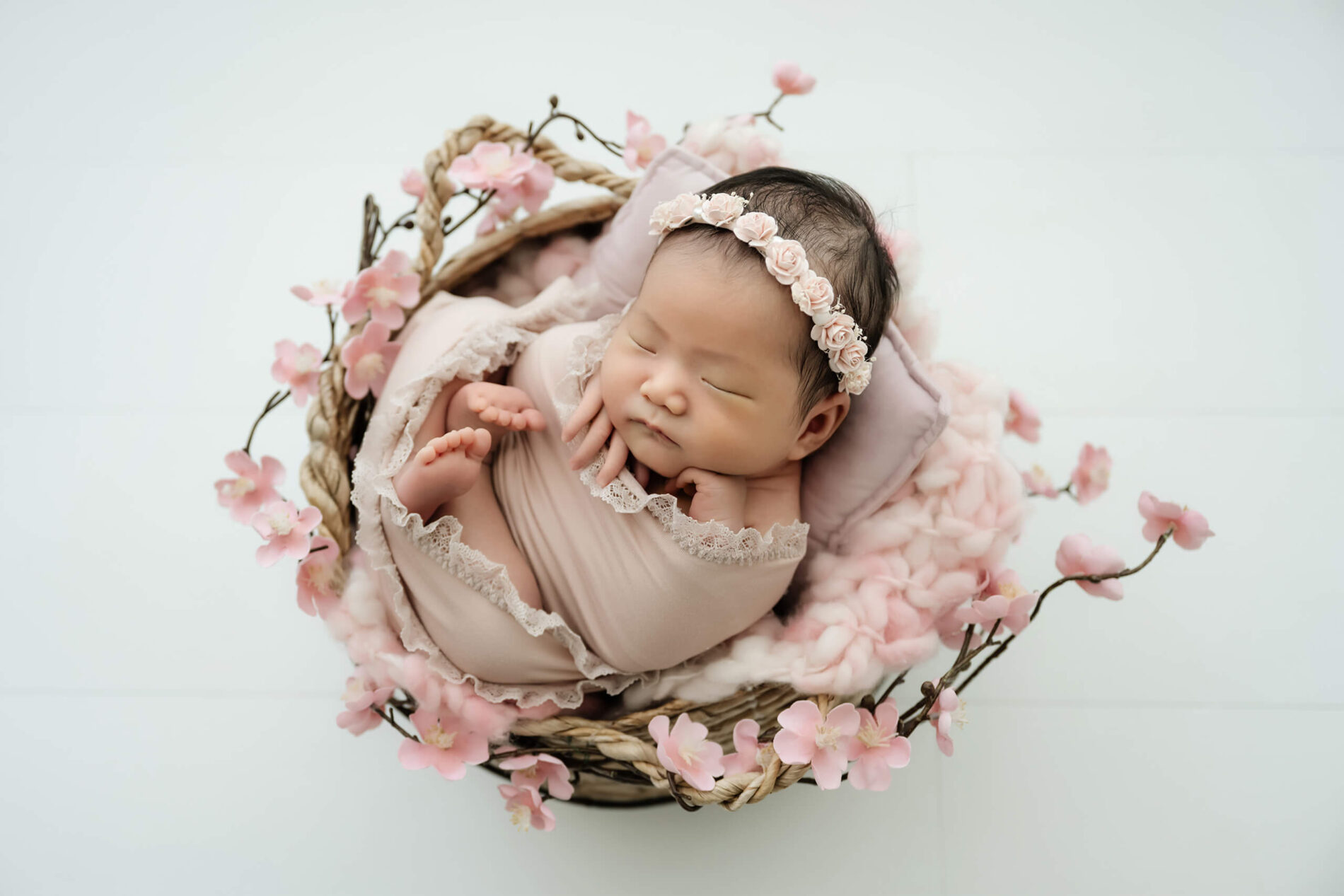 Overhead portrait of a newborn wrapped in soft blush tones, resting in a basket with delicate pink florals during a Seattle studio newborn session.