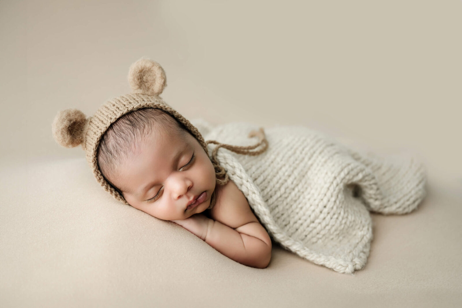 Sleeping newborn photographed in a Seattle studio, wearing a neutral knit bonnet and wrap, posed on a soft beige backdrop during a newborn photoshoot