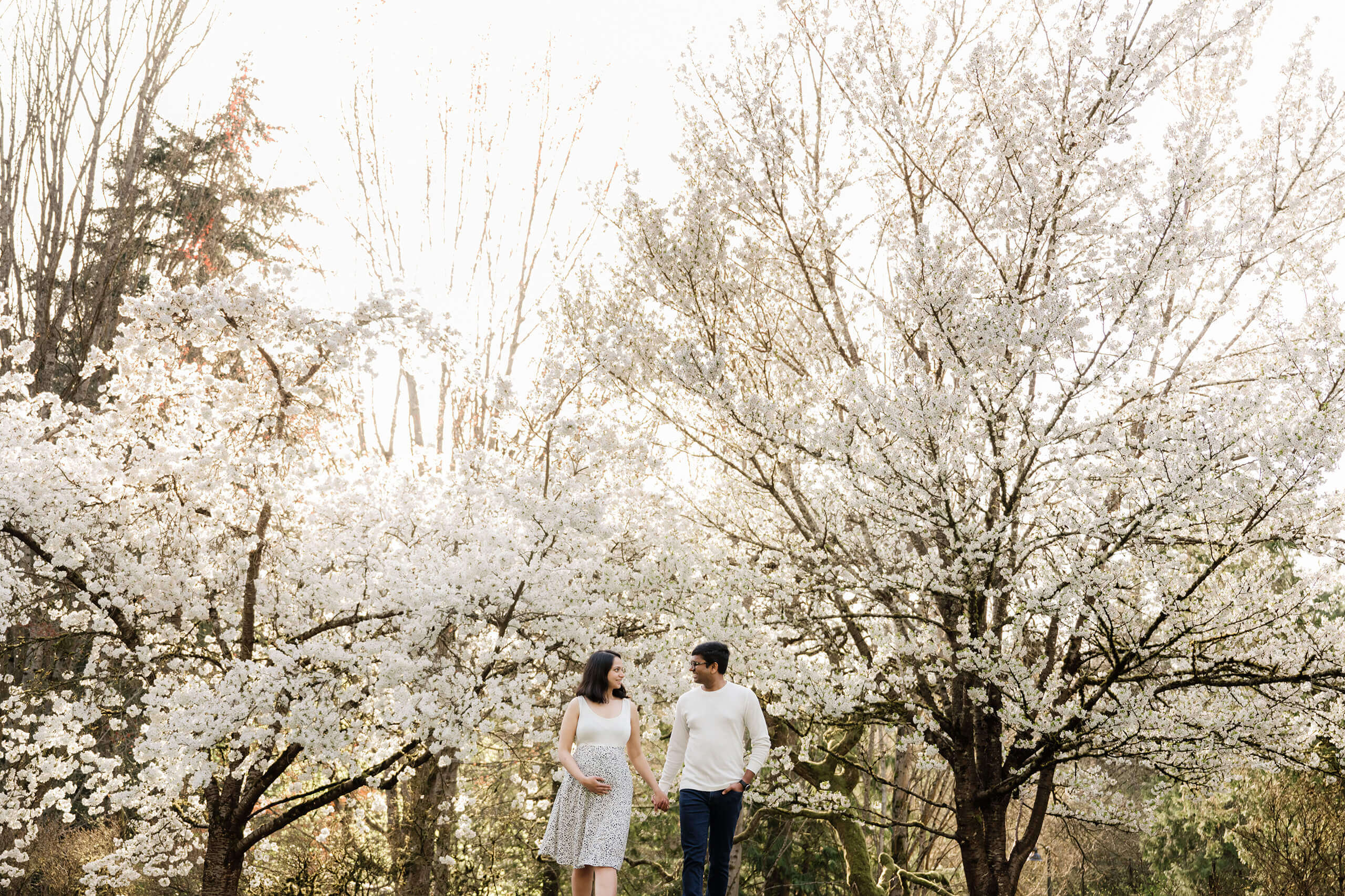 Expecting couple walks hand in hand beneath white cherry blossoms in Seattle, light and airy maternity portrait.