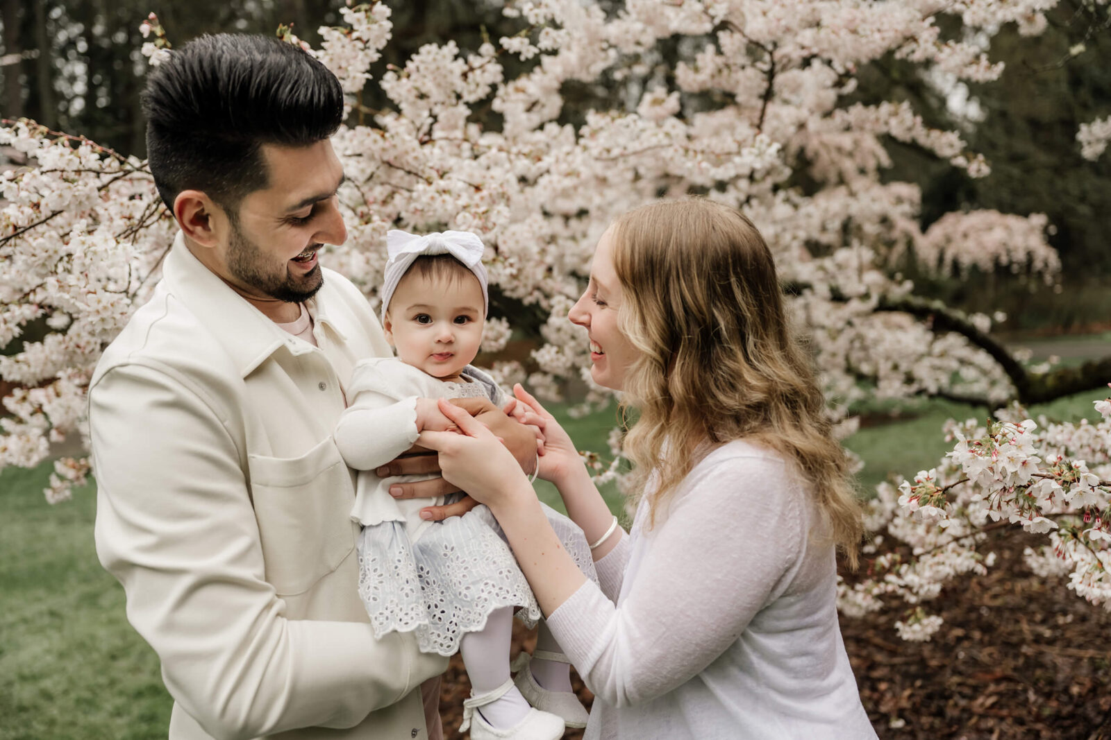 Parents smile at their baby in front of pale pink cherry blossoms, close-up Seattle spring family portrait.