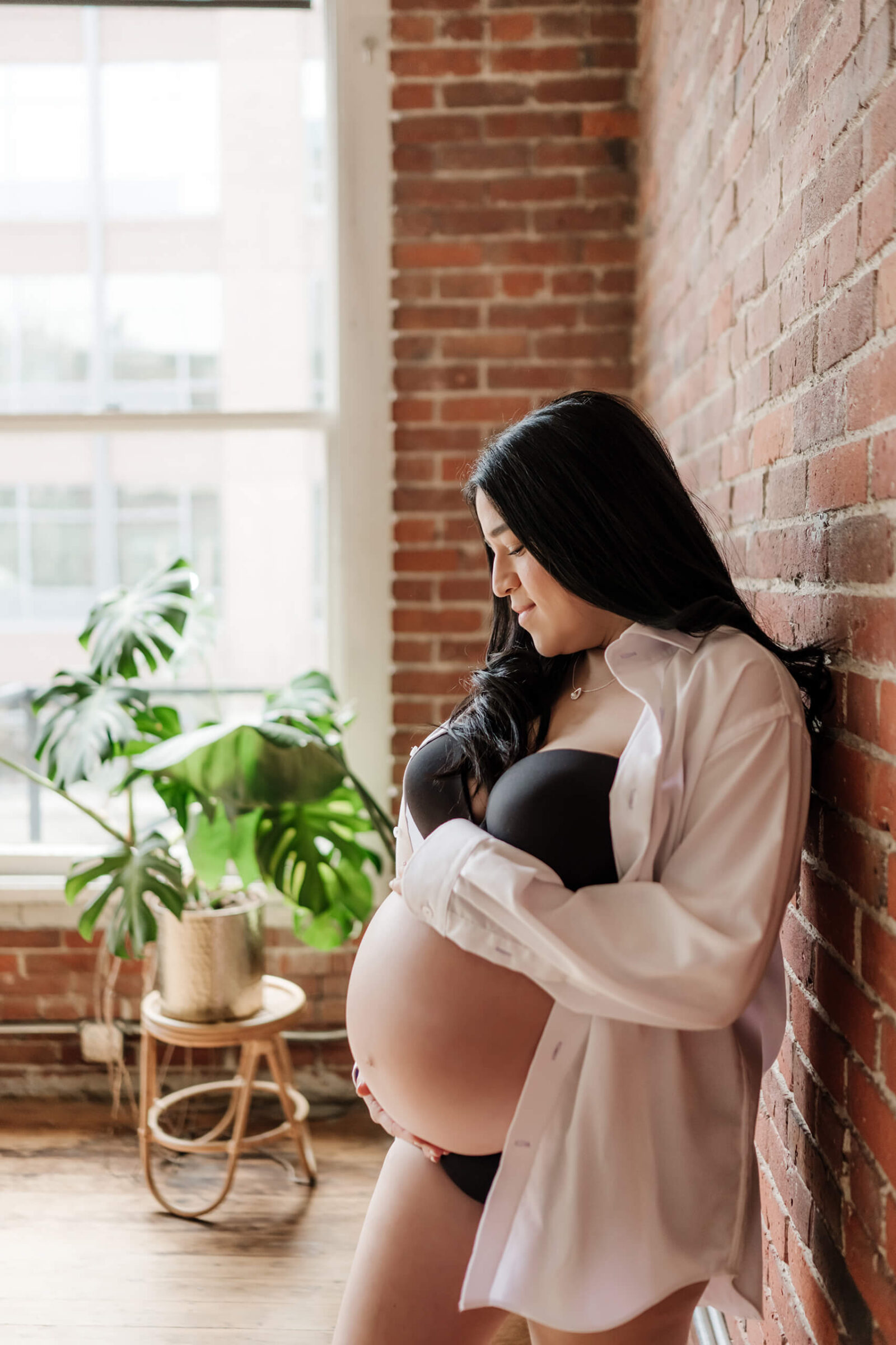 Pregnant woman in a black bra and open white shirt, leaning against a brick wall by a bright studio window with a leafy plant nearby.