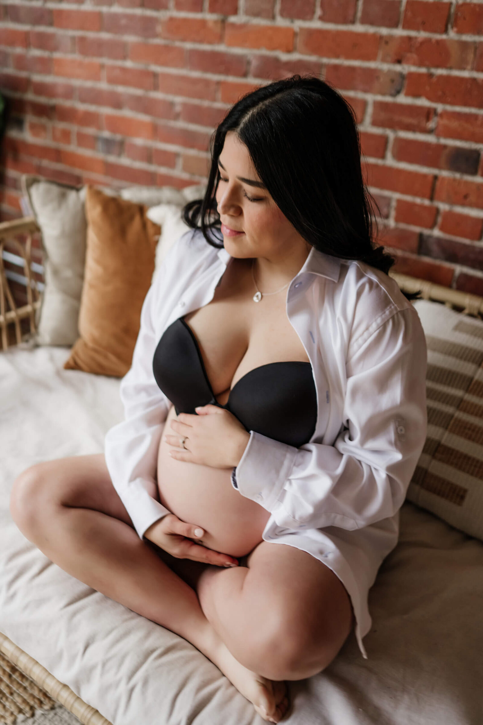 Maternity portrait on a bed in a brick-walled studio; mom-to-be sits cross-legged in a black bra and open white shirt, hands resting on her belly.