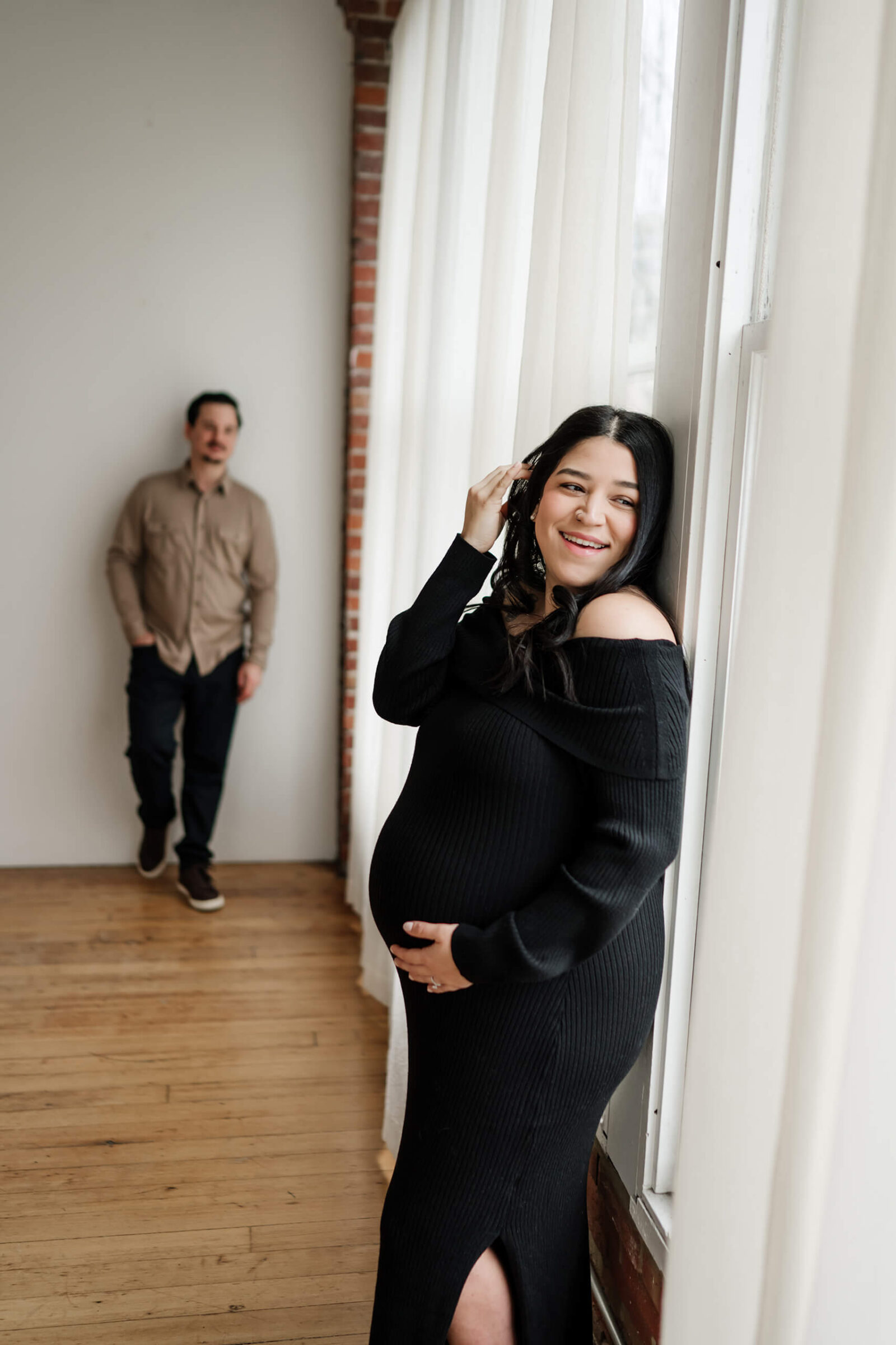 Pregnant woman in a black off-the-shoulder dress smiling by a tall studio window with white curtains, with her partner softly blurred in the background.