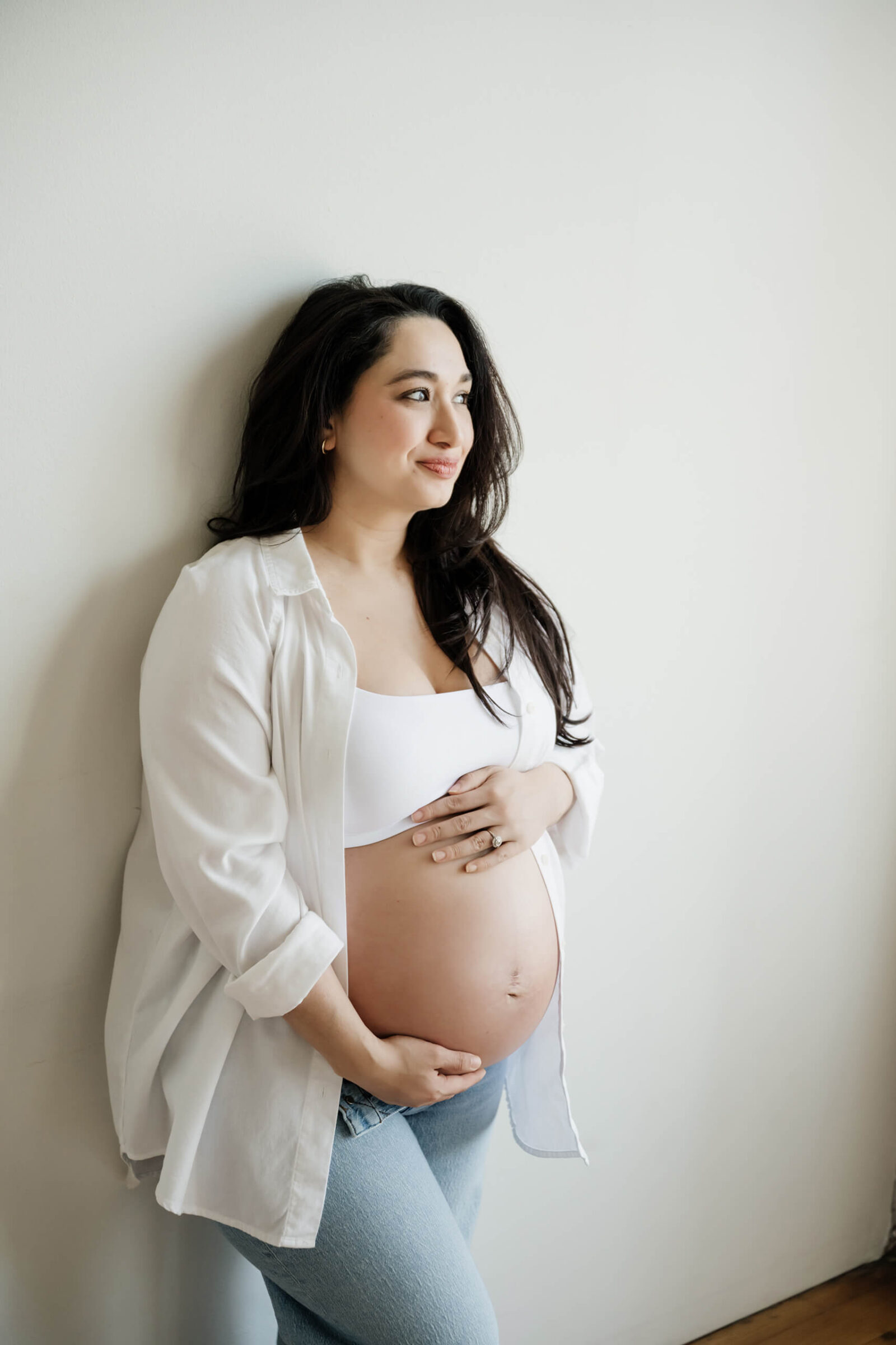 Pregnant mom-to-be in a white shirt and jeans leaning against a simple studio wall, hands on her baby bump