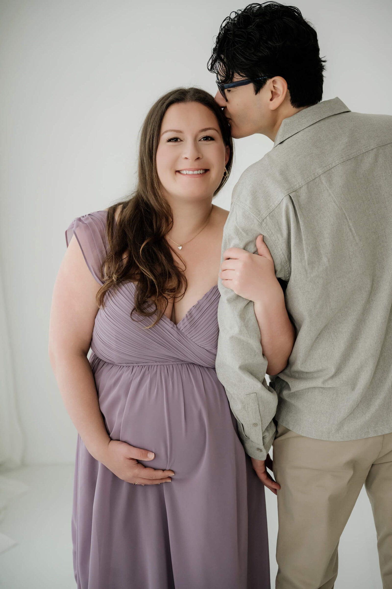 Couple maternity portrait in a white studio, partner kissing her forehead as she smiles in a lavender dress.