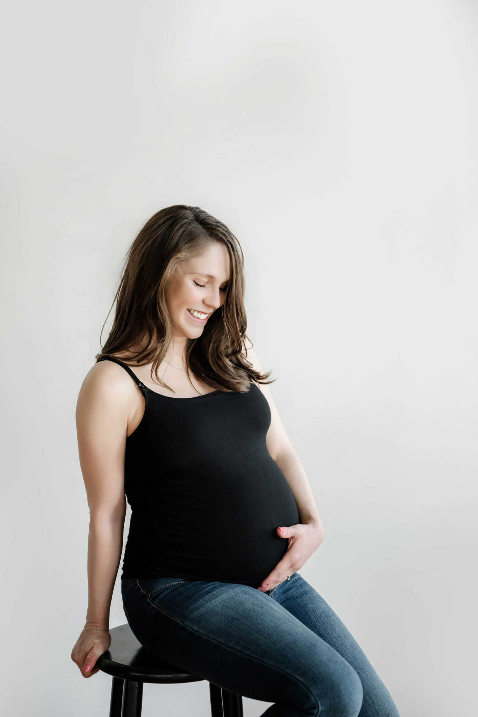 Seated studio maternity portrait of an expecting mother in a black tank top and jeans, smiling down at her belly.