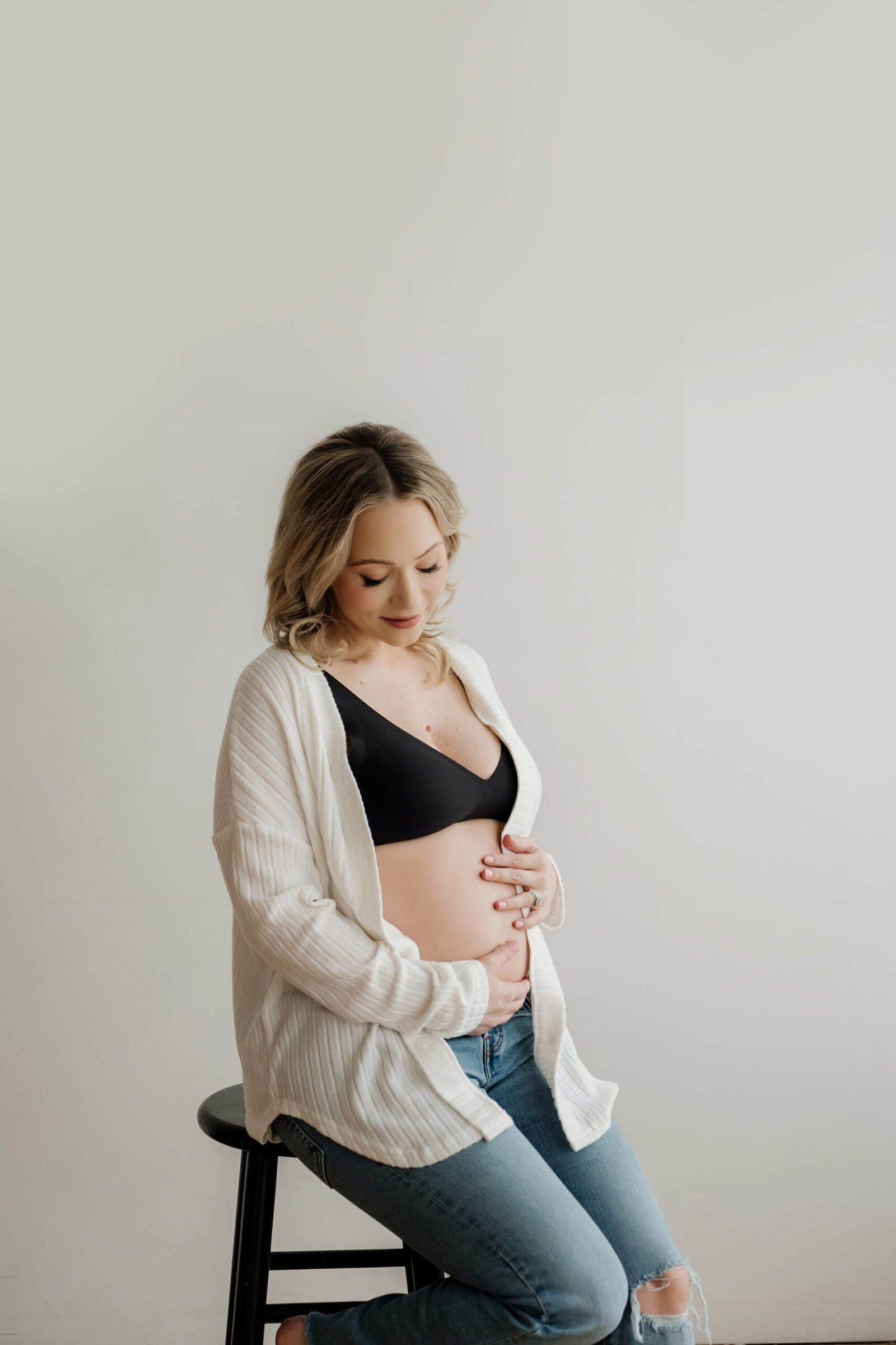 Pregnant woman seated on a stool in jeans, black bra, and white cardigan, gently holding her belly in a simple white studio.