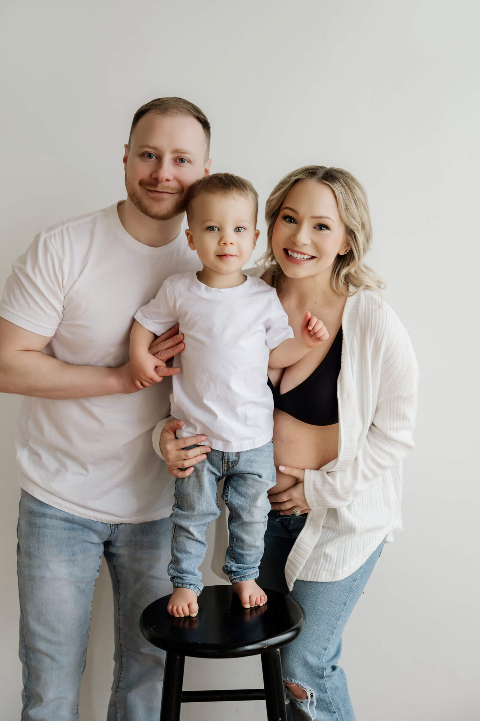 Studio family maternity portrait with parents holding their toddler on a black stool against a simple white wall.