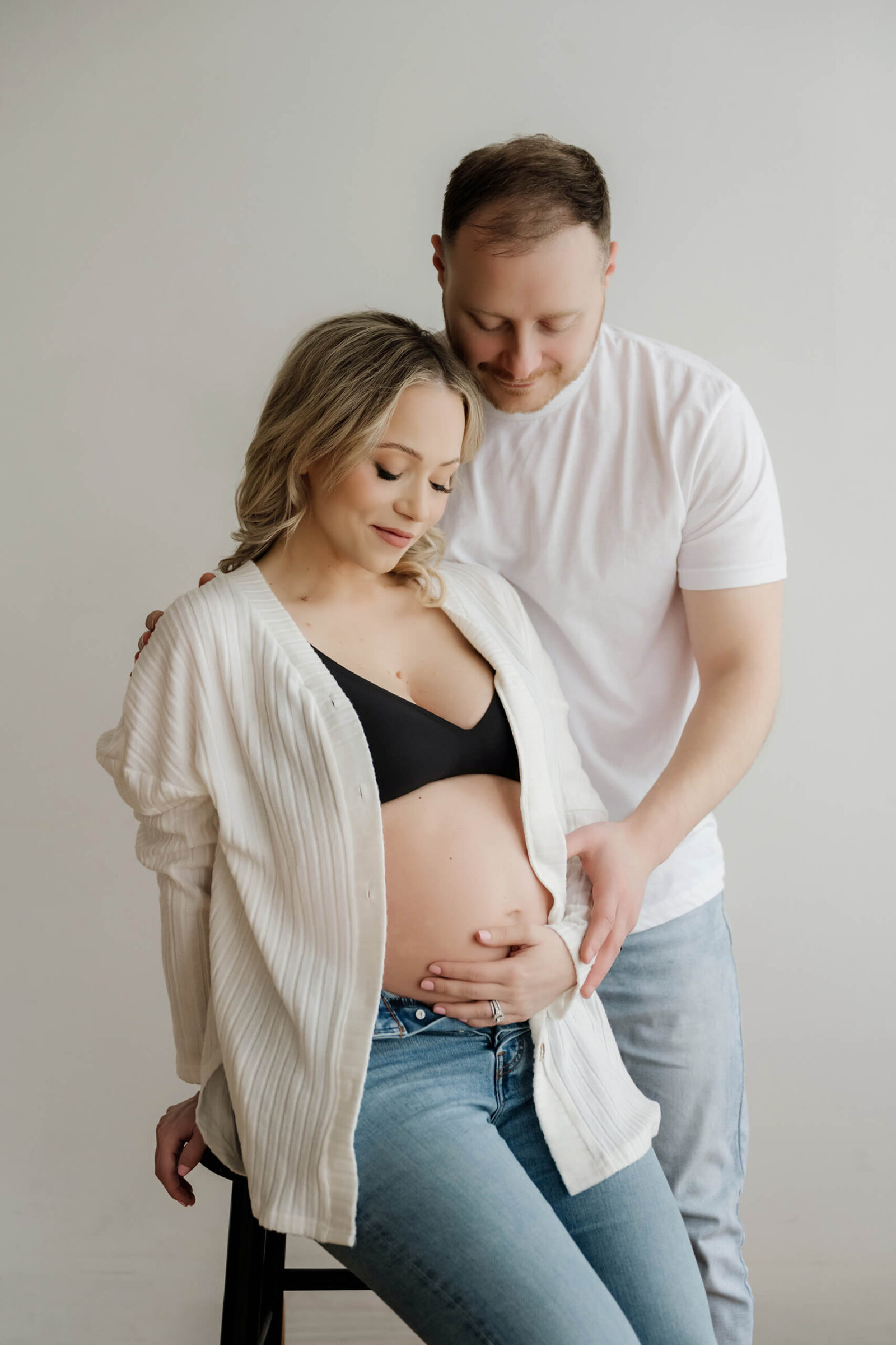 Couple maternity portrait in a white studio, expecting mother seated in jeans and cardigan while partner stands behind with hands on her belly.