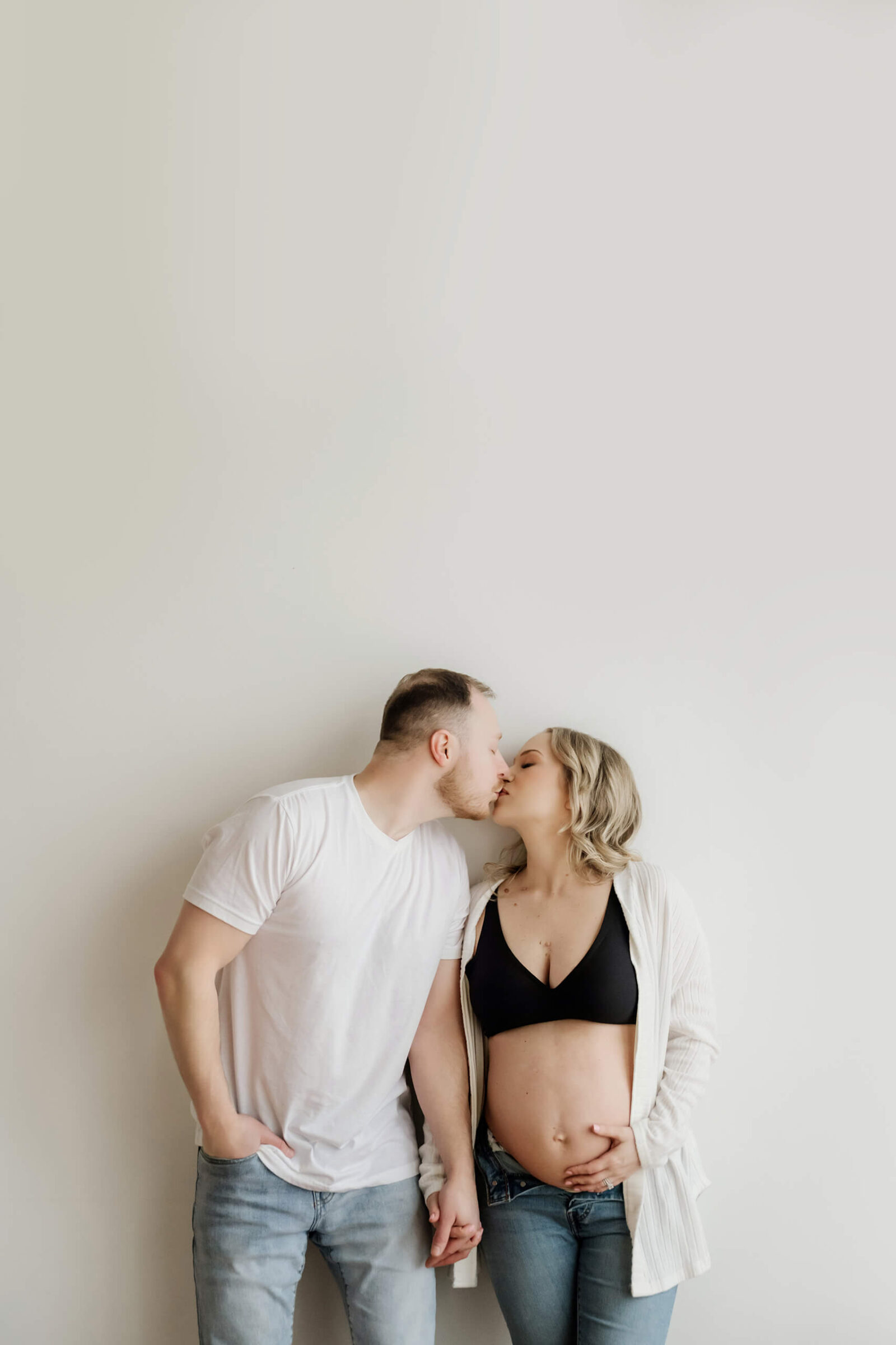 Couple maternity portrait in a white studio, partners holding hands and kissing while she cradles her bare belly.