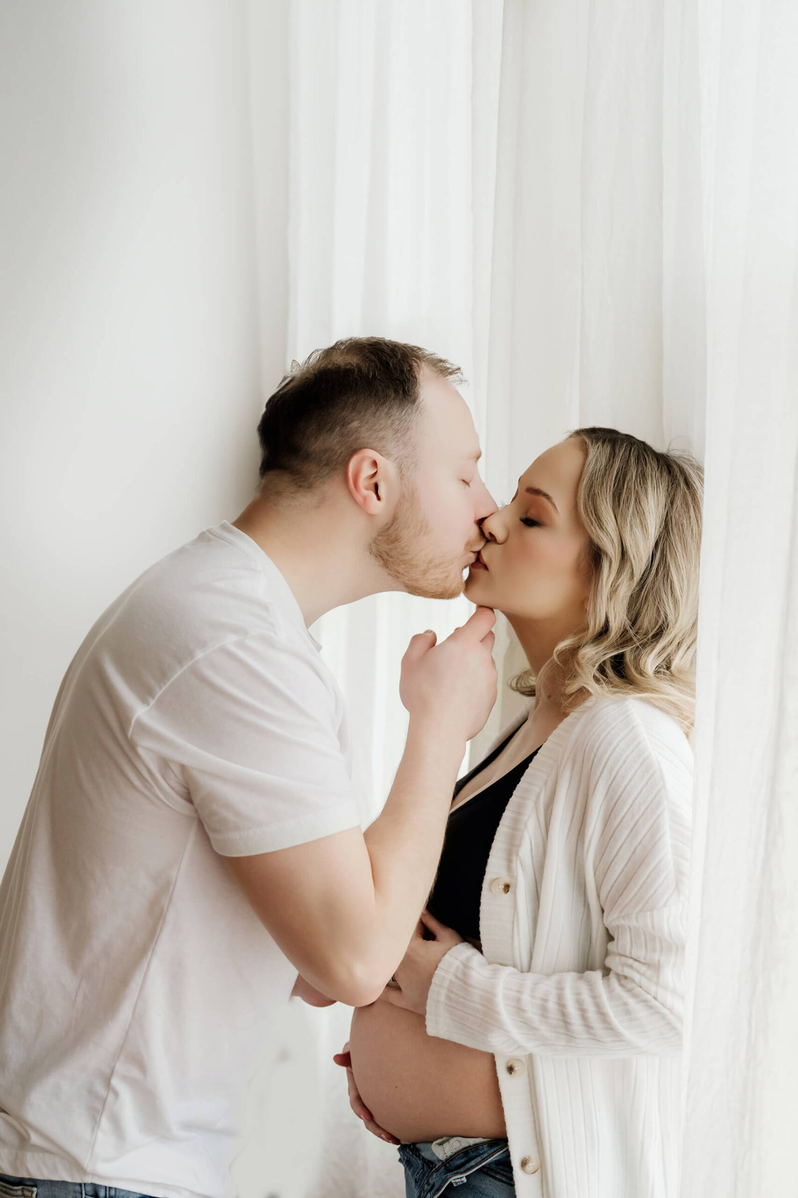 Close-up maternity portrait by a bright studio window, couple sharing a kiss as she rests a hand on her belly.