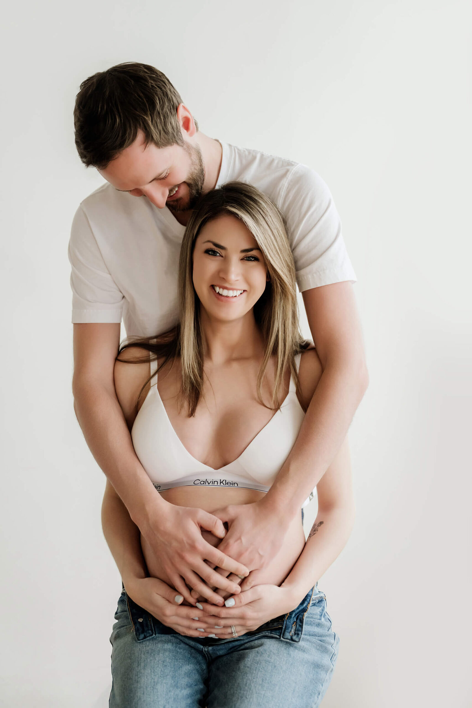 Expecting couple in white tops and jeans, standing against a clean white studio wall with hands gently holding the baby bump.