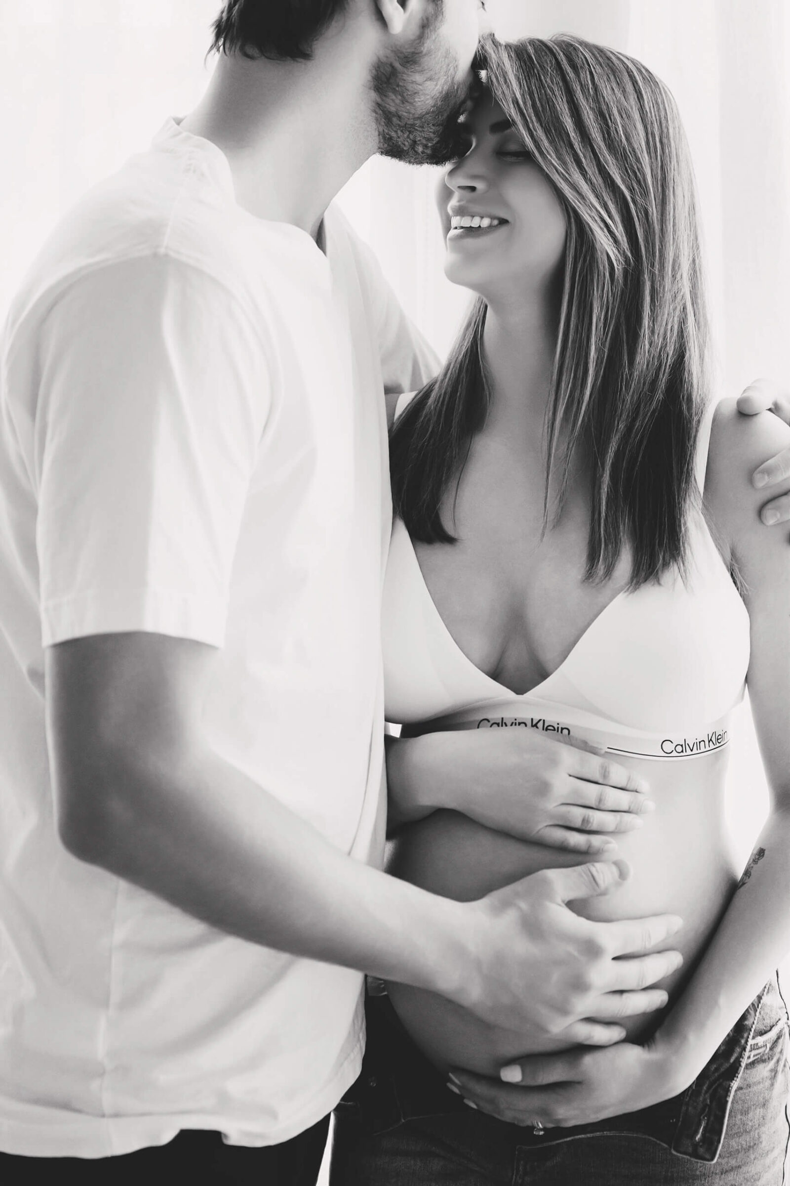 Black-and-white maternity portrait of an expecting couple by the window as the dad kisses mom’s forehead, both smiling.