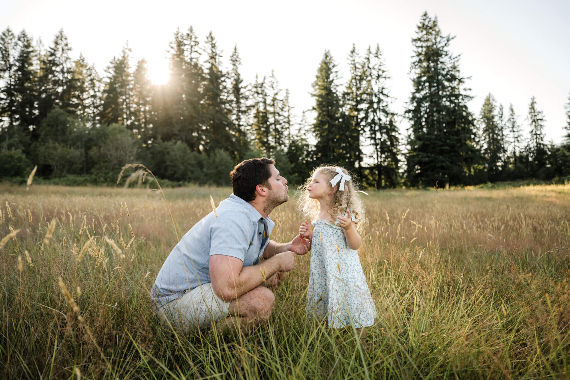 Father and daughter sharing a playful moment in a grassy field with evergreen trees behind them.