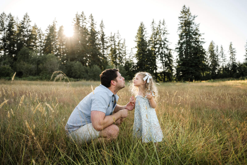 Father and daughter sharing a playful moment in a grassy field with evergreen trees behind them.
