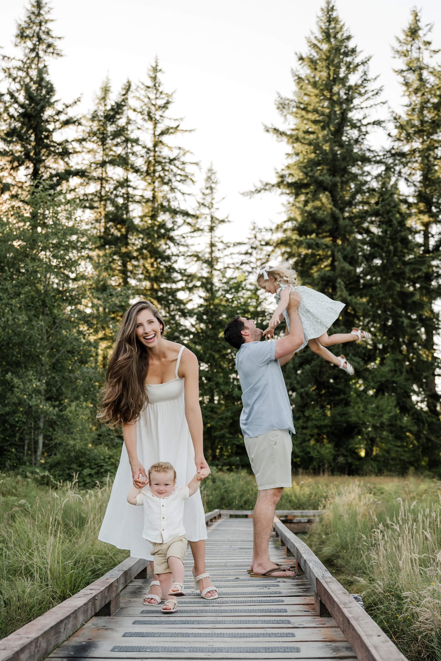 Family of four on a wooden boardwalk, with one parent helping a toddler walk while the other lifts a child in the background.