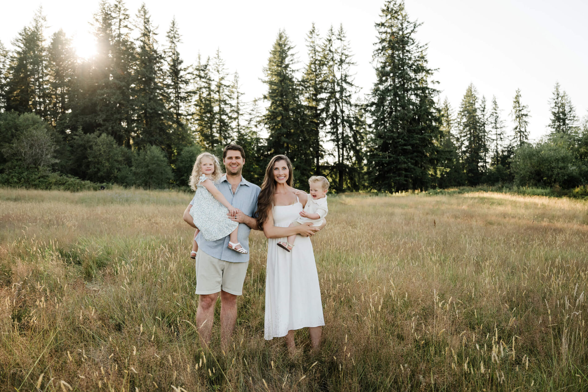 Family of four standing together in a tall golden field with evergreen trees behind them.