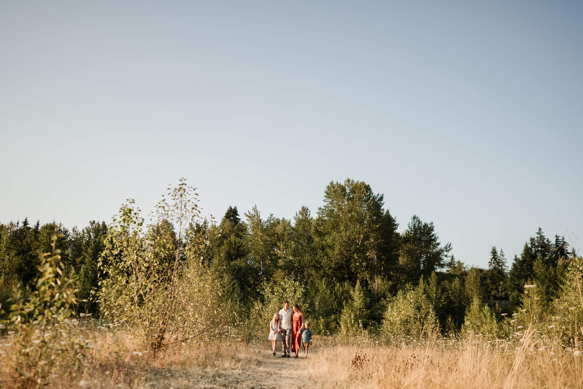 Family of four walking together through a golden field with trees and open sky in the background.