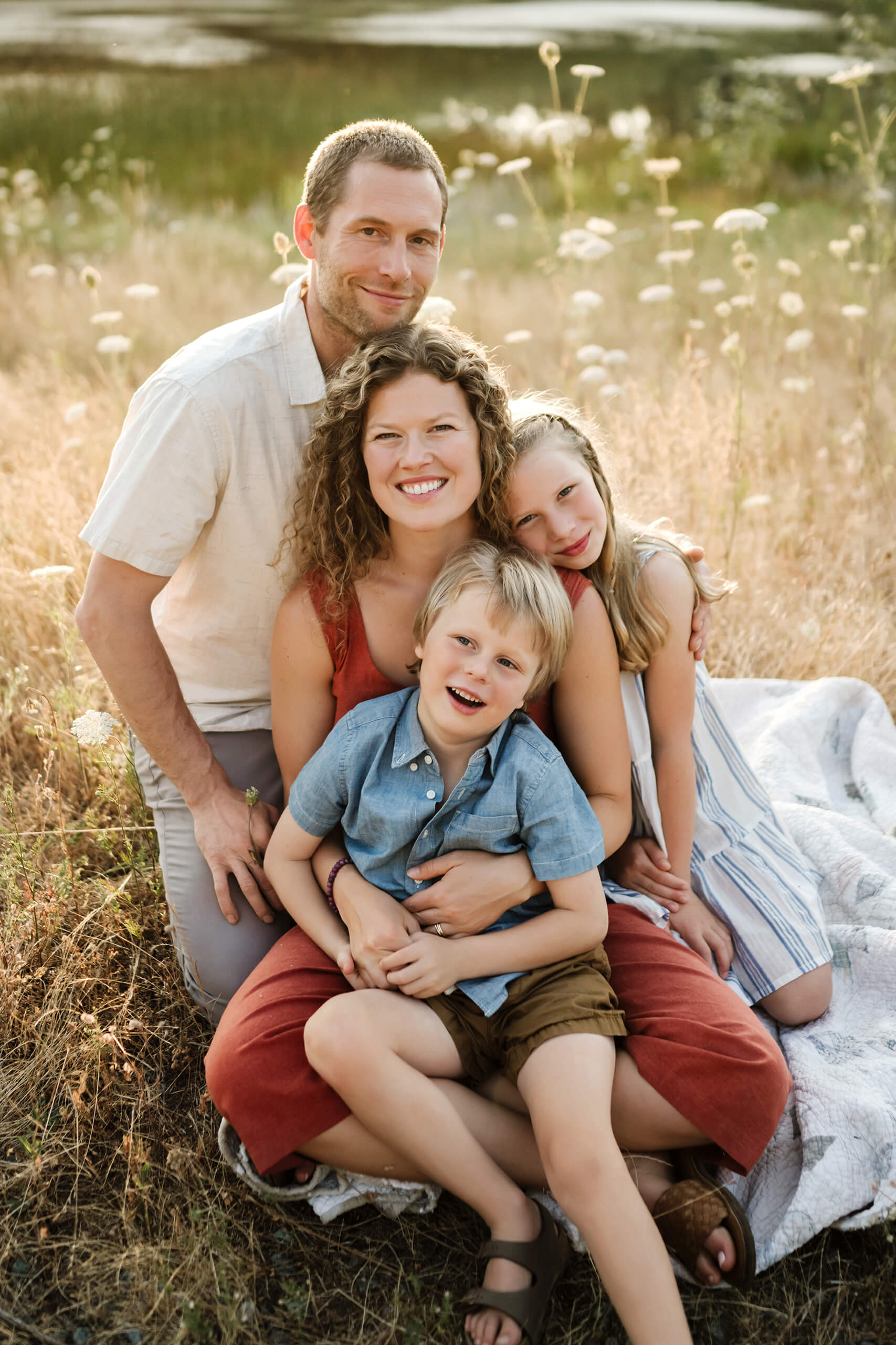 Family of four cuddling together on a quilt in a golden field near the water.