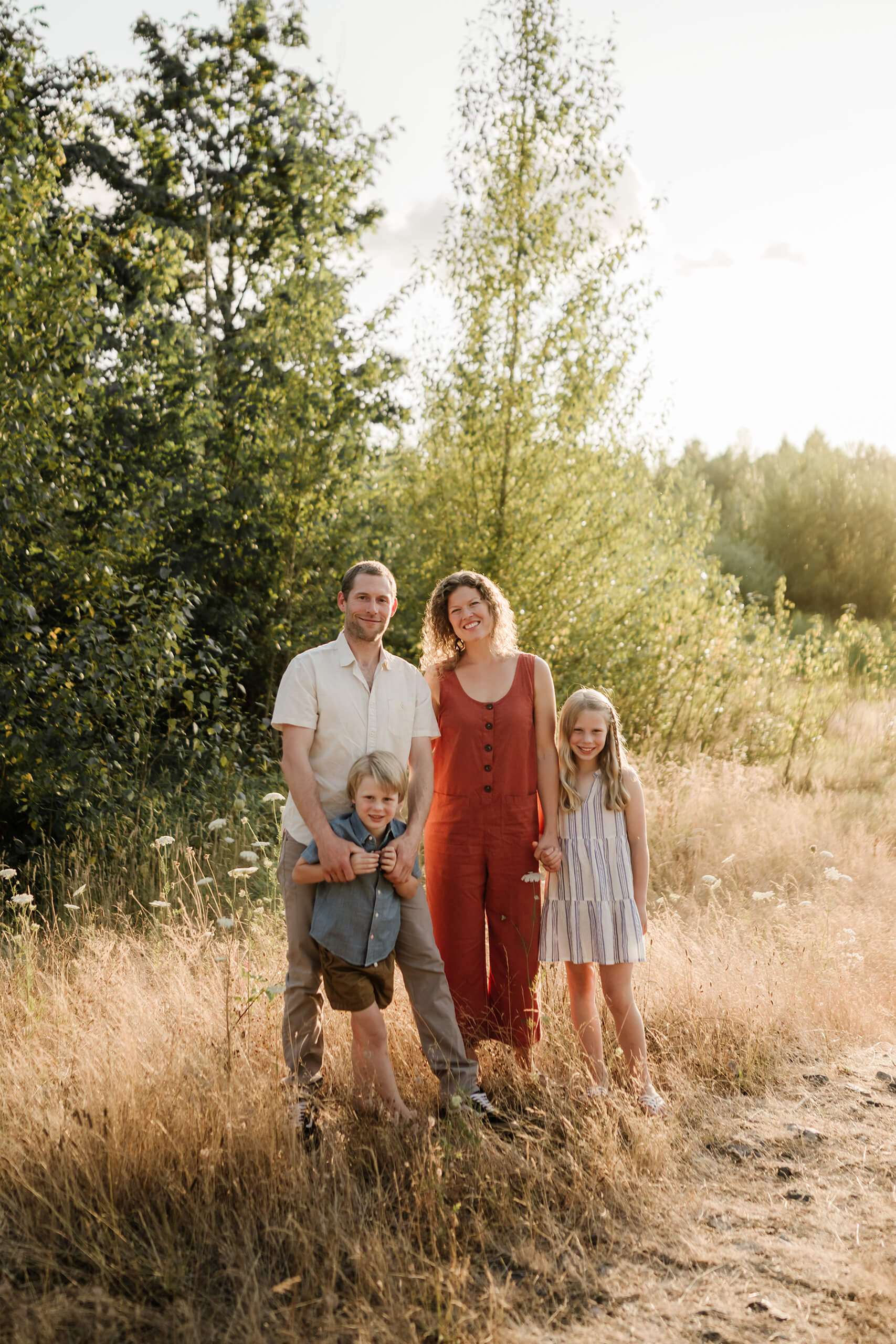 Family of four standing together in tall dry grass with trees and soft evening light behind them.
