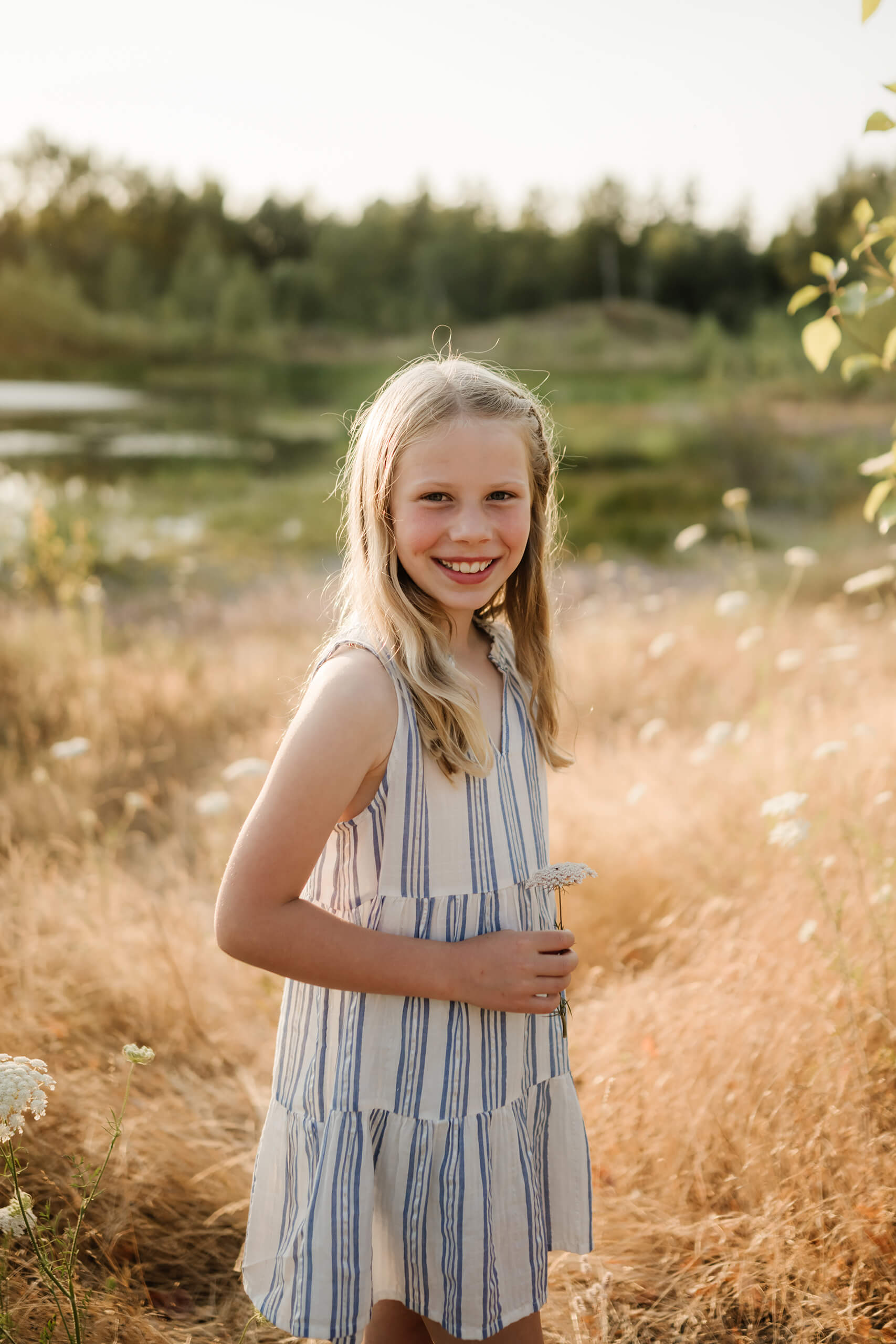 Young girl smiling in a golden field near a pond while holding a small white flower.