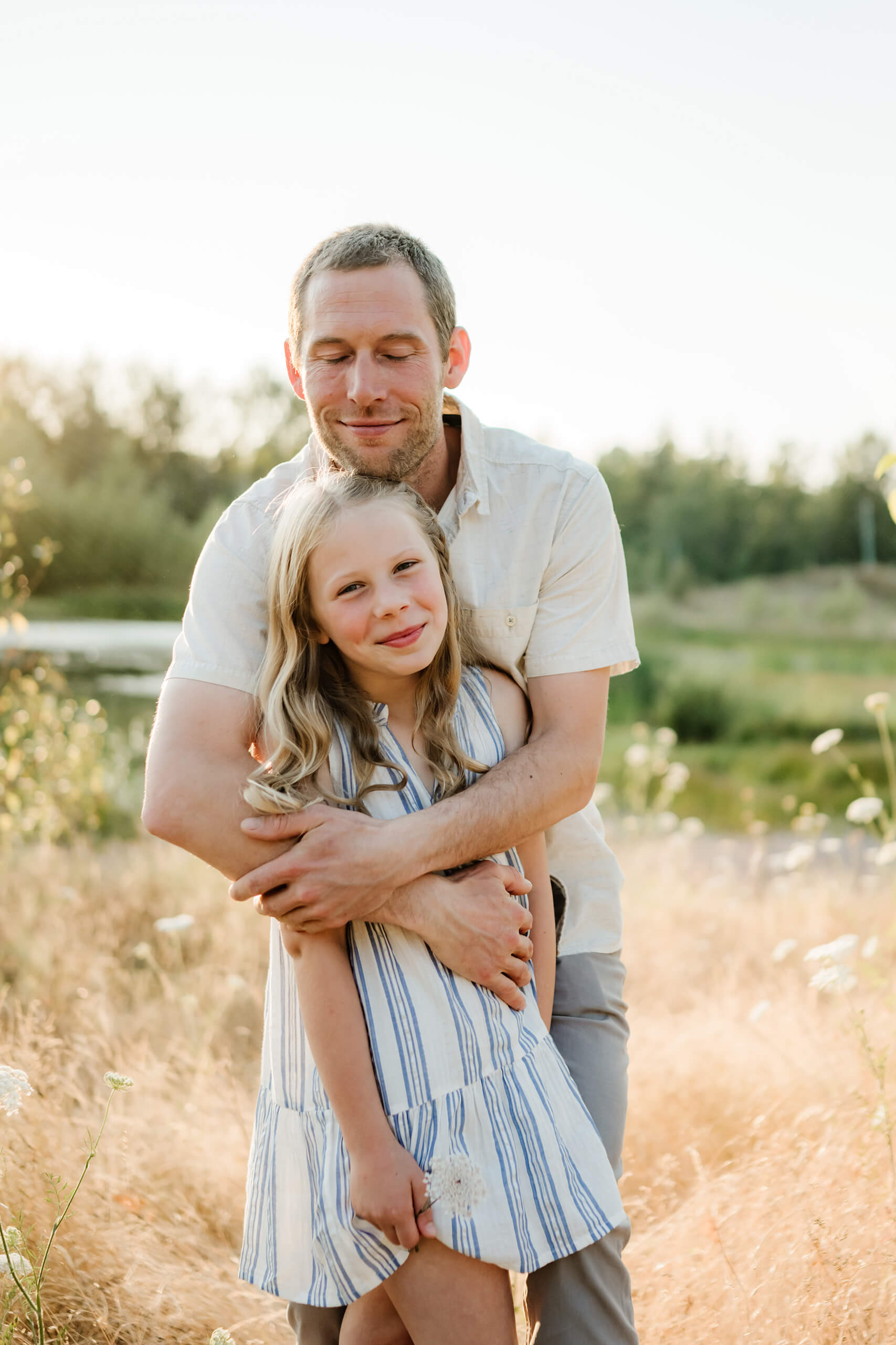 Father hugging his daughter in a sunlit field near the water during an outdoor family session.