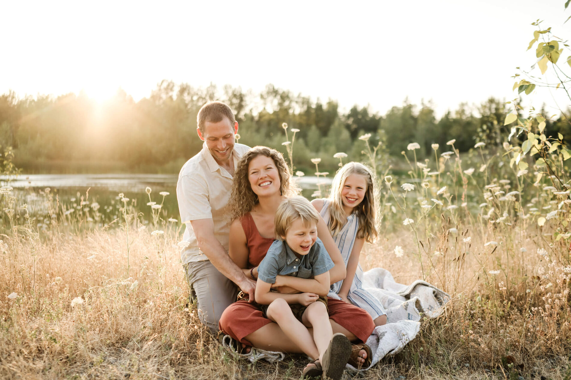 Family of four sitting together on a quilt in golden grass near the water.