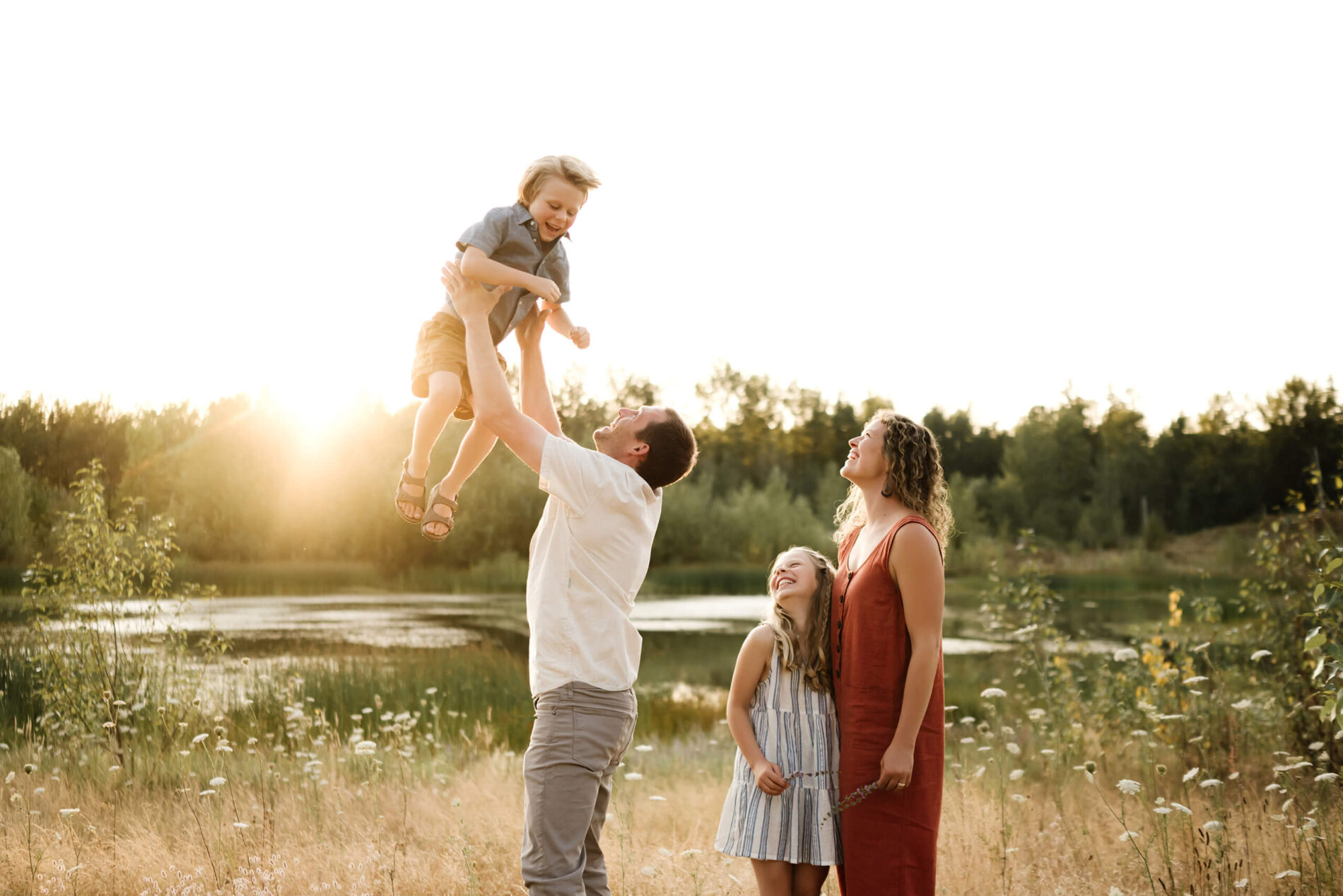 Father lifting his son in a sunlit field while mother and daughter watch nearby.
