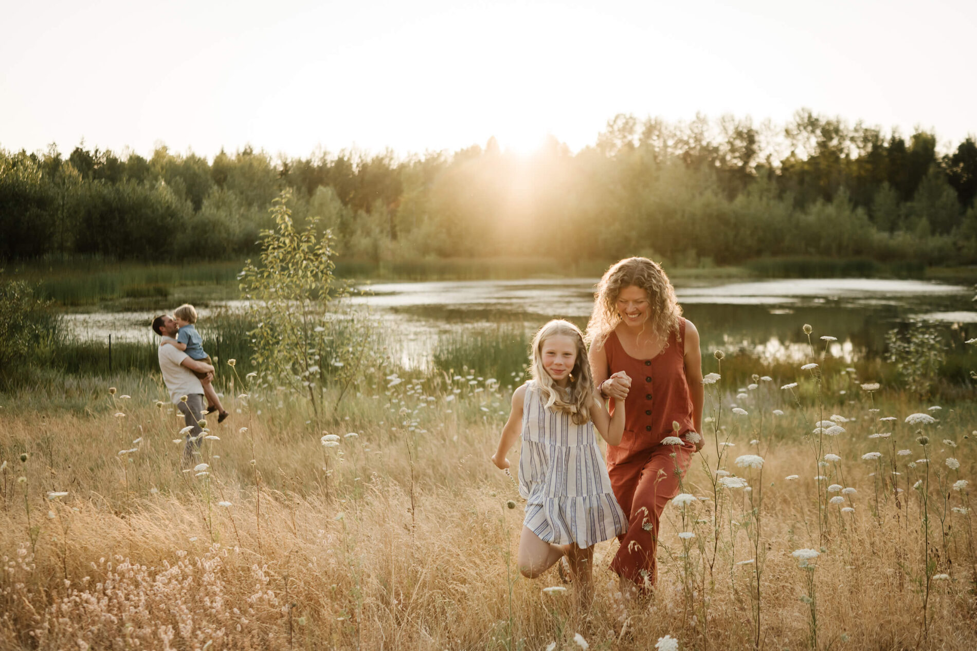 Mother and daughter walking through golden grass near a pond, with father and son in the background.