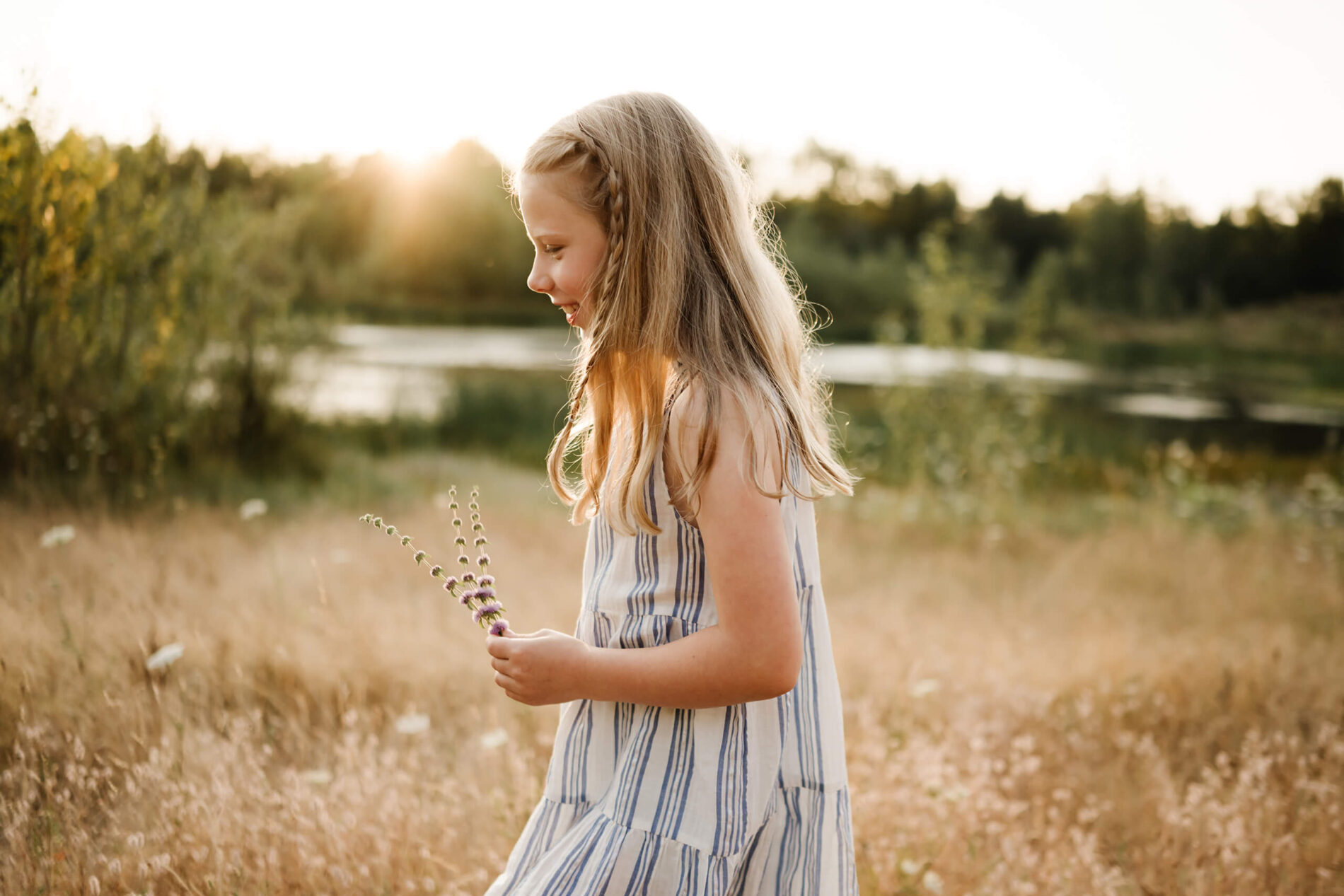 Young girl looking down at purple wildflowers in a sunlit field near a pond.