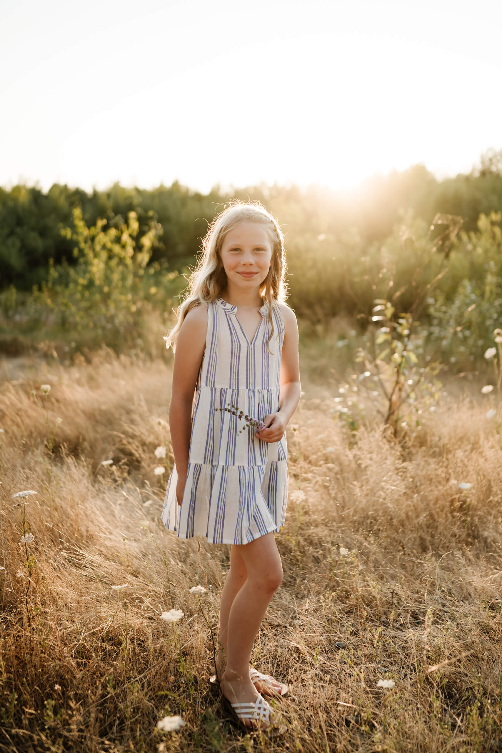Young girl standing in tall golden grass while holding a small wildflower near the water.
