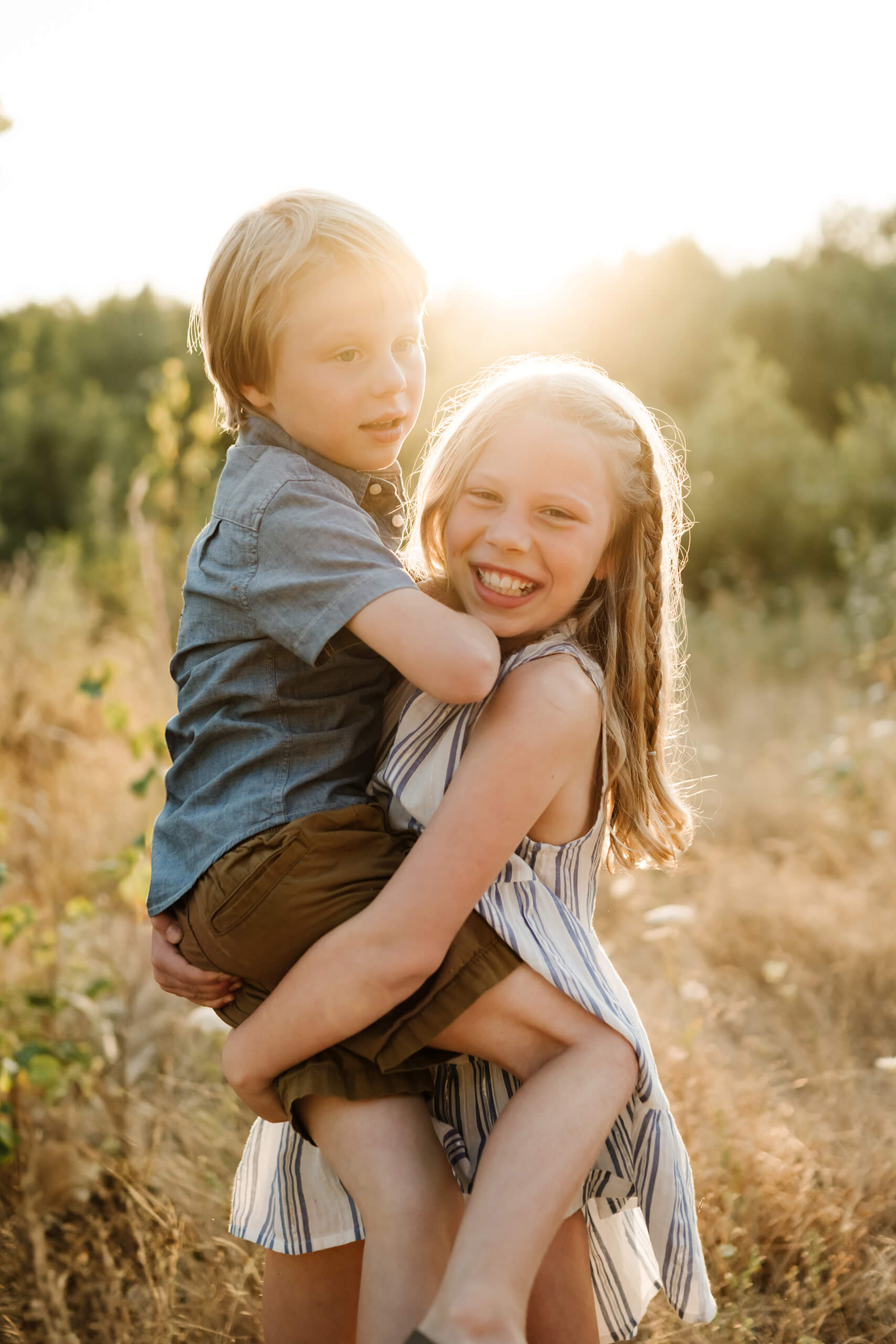 Young girl carrying her little brother in a golden field during an outdoor family photo session.