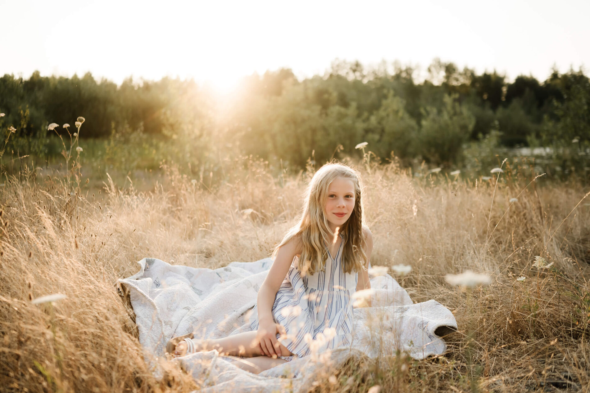 Young girl sitting on a quilt in a sunlit field with tall golden grass around her.