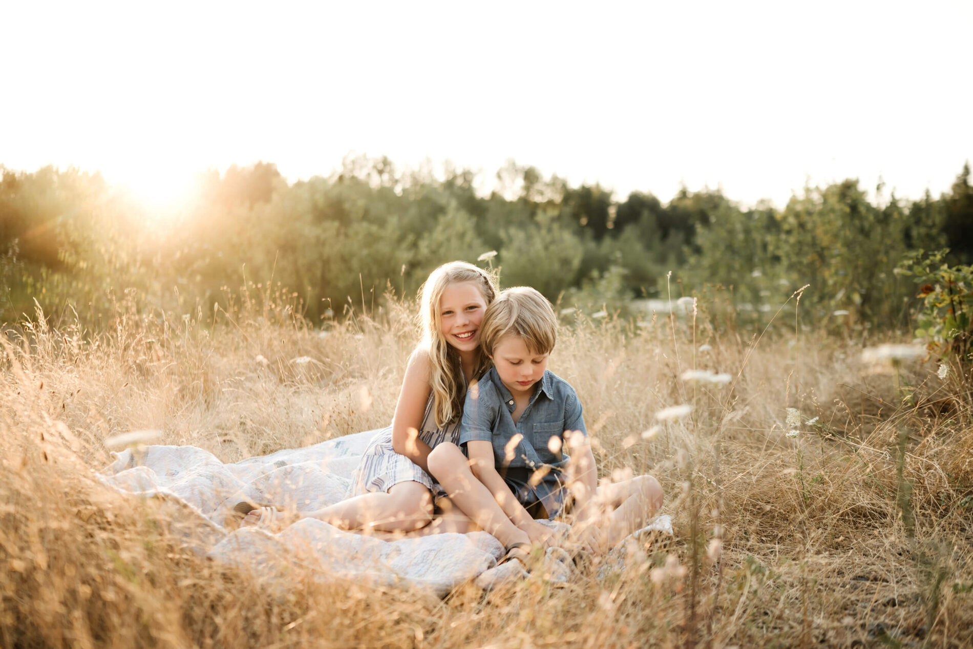 Brother and sister sitting together on a quilt in tall grass during golden hour.