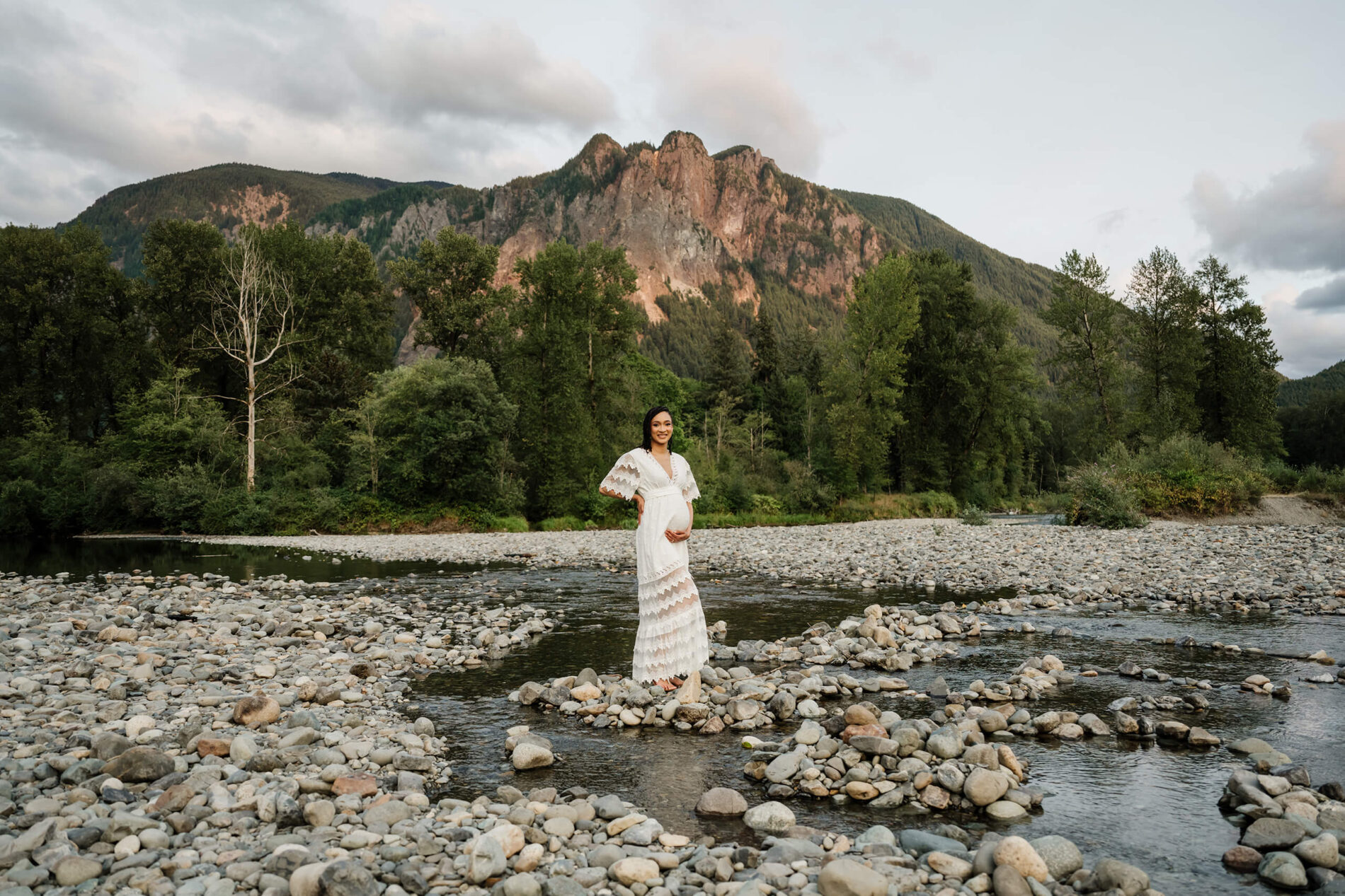 Pregnant woman in a white lace dress standing on a rocky riverbank with forest and mountains behind her.