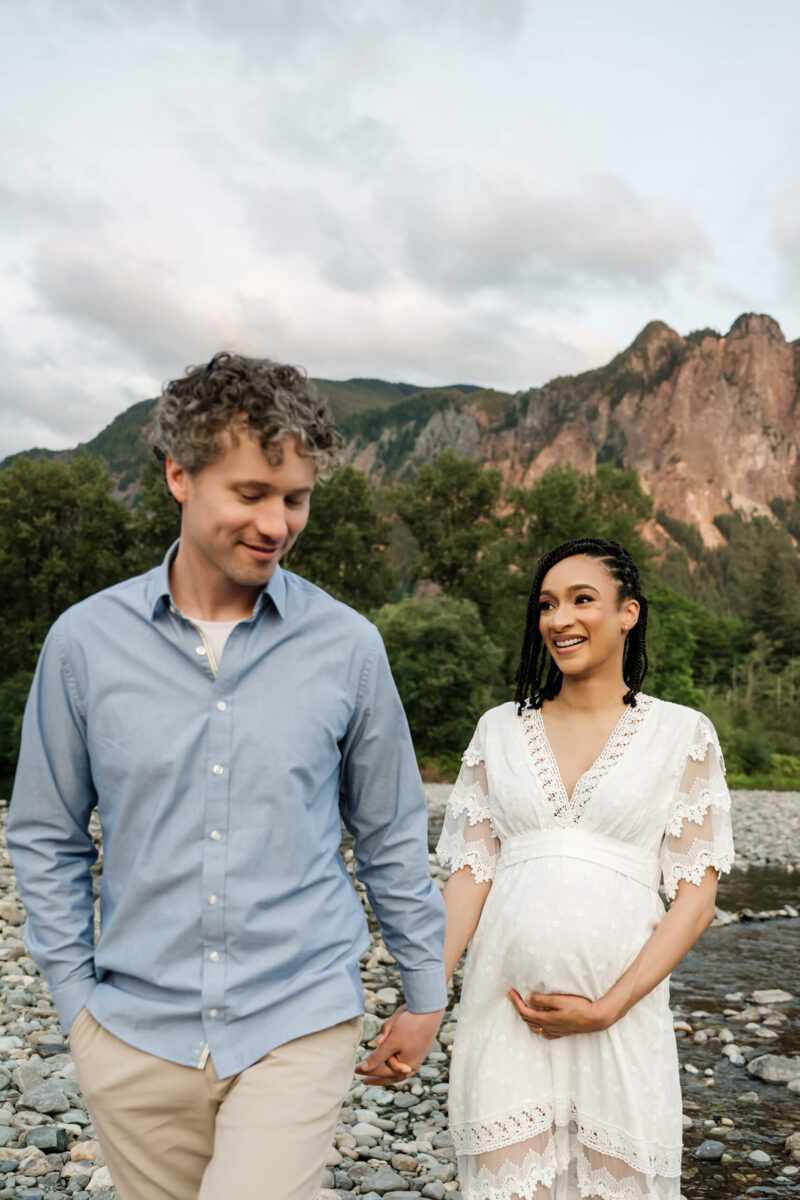 Expecting couple holding hands during an outdoor maternity session with mountains and a rocky river in the background.