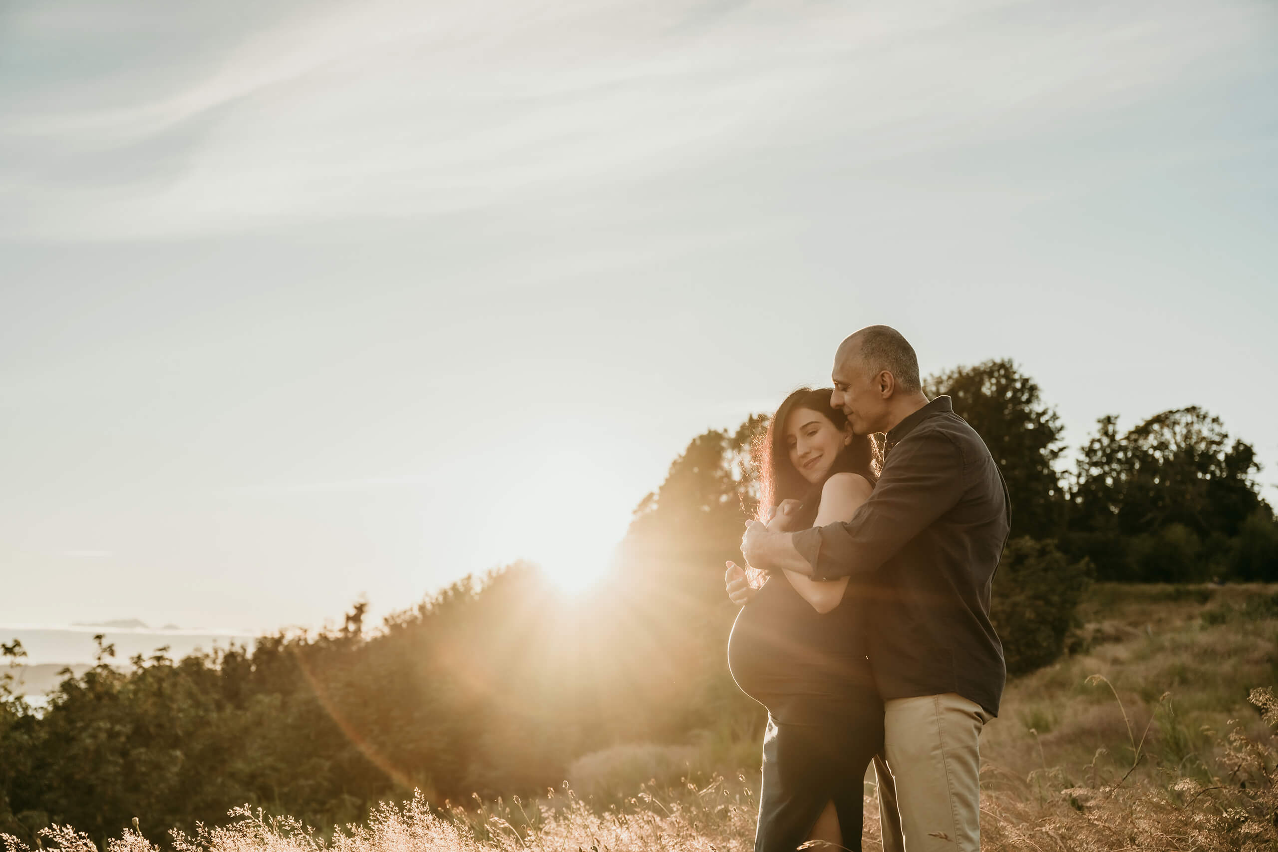 Expecting couple embracing in a sunlit field during an outdoor maternity session.