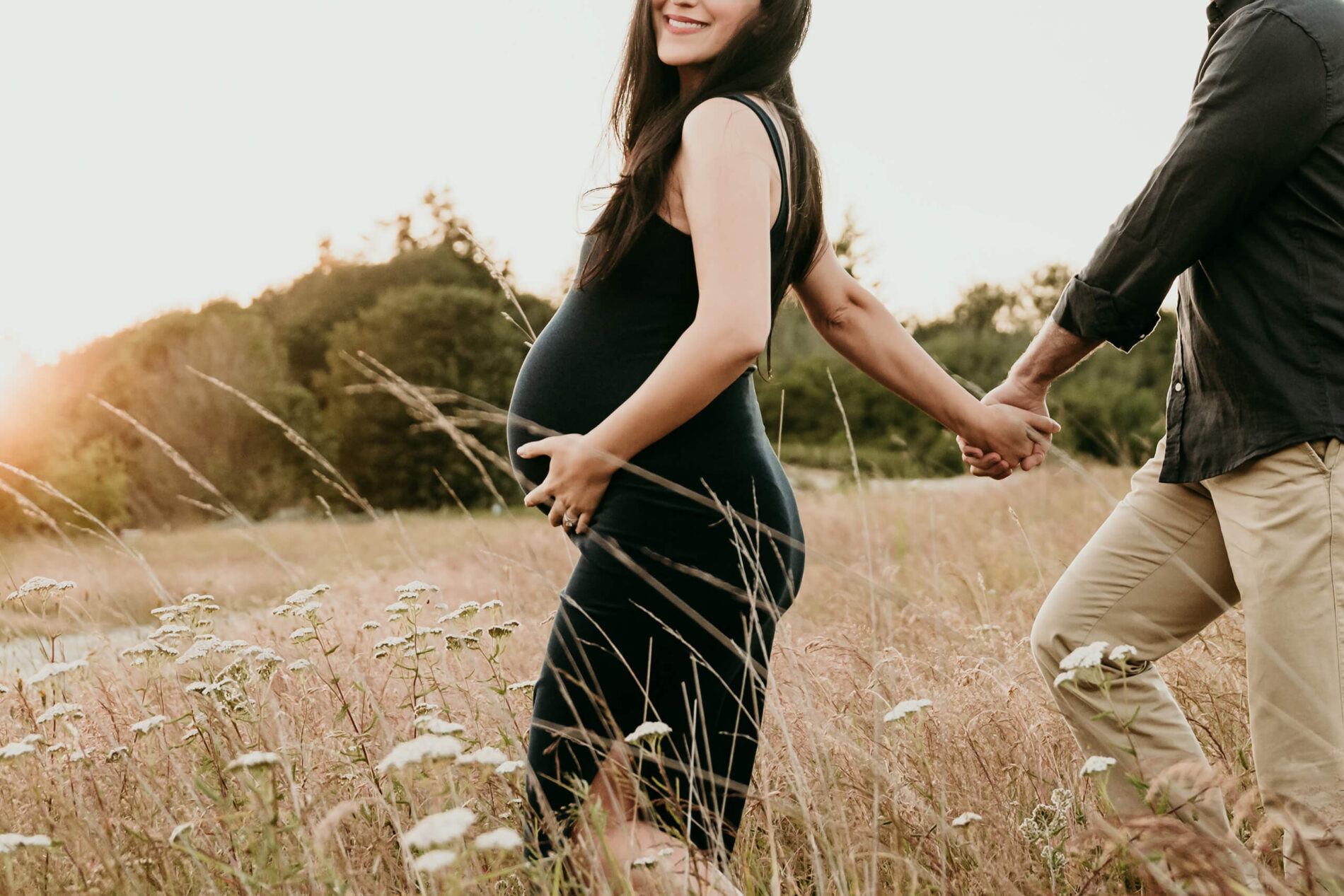 Pregnant woman in a black dress holding hands with her partner while walking through a grassy field.