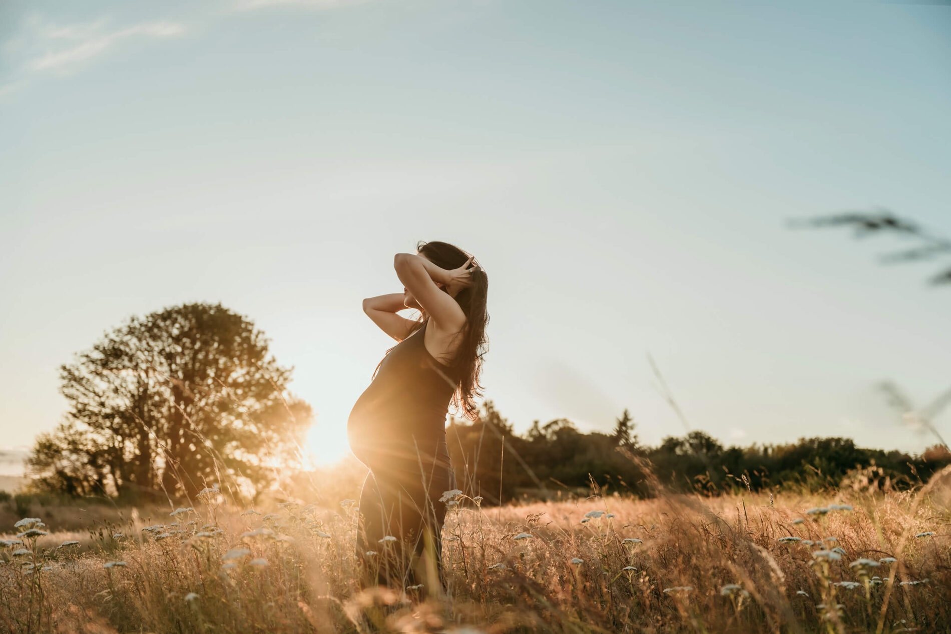 Pregnant woman standing in tall grass at sunset for a warm outdoor maternity portrait.
