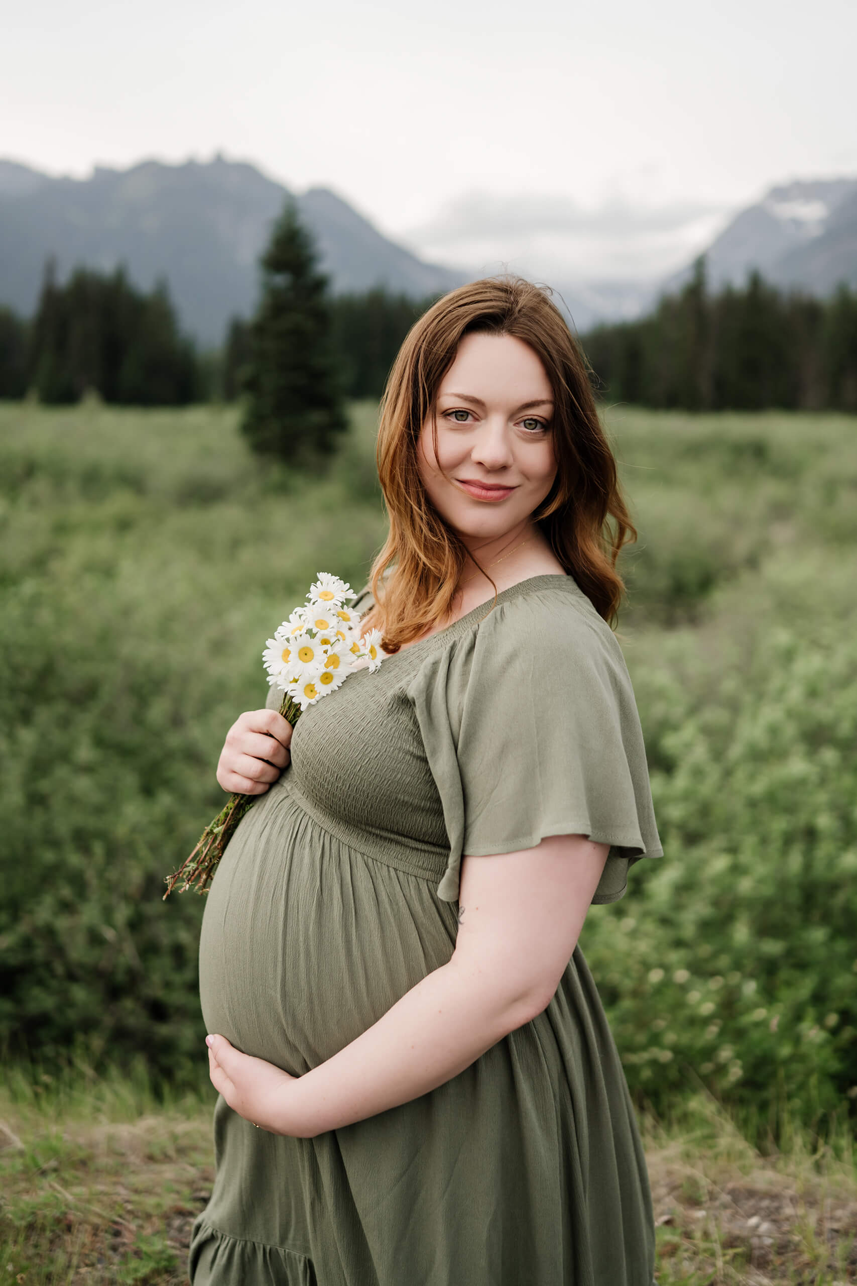 Pregnant woman holding white flowers in a green meadow with forested mountains behind her.