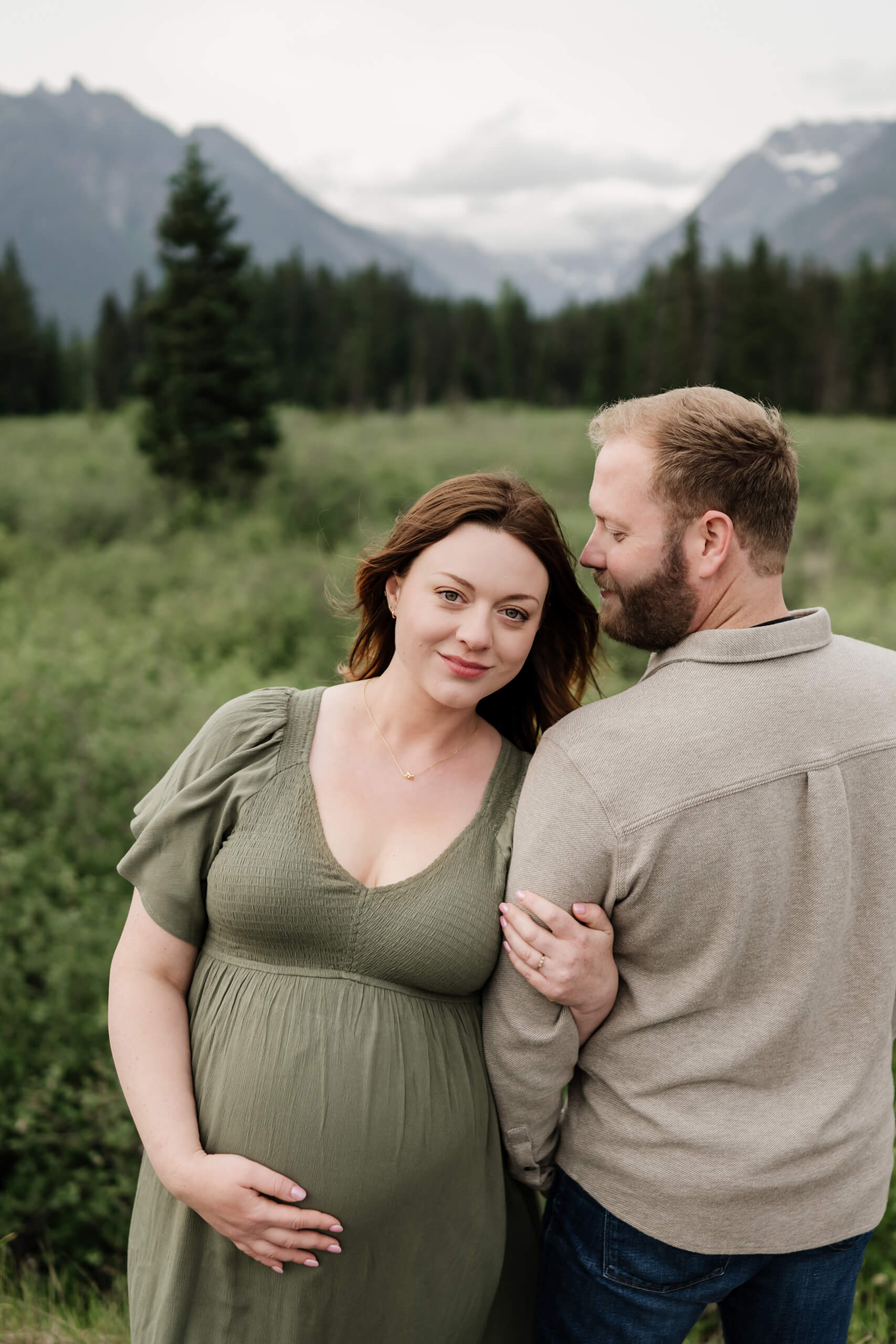 Pregnant woman in a green dress standing with her partner in a mountain meadow.