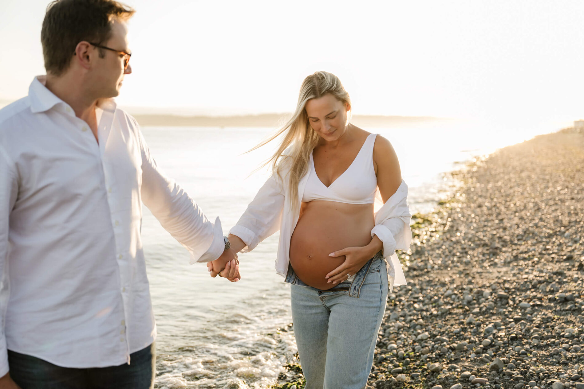 Expecting couple holding hands while walking along a rocky beach during a sunset maternity session.