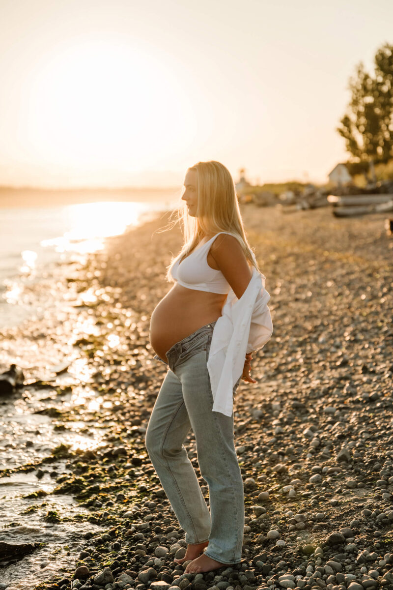 Pregnant woman in jeans and a white top standing in profile on a rocky beach at sunset.