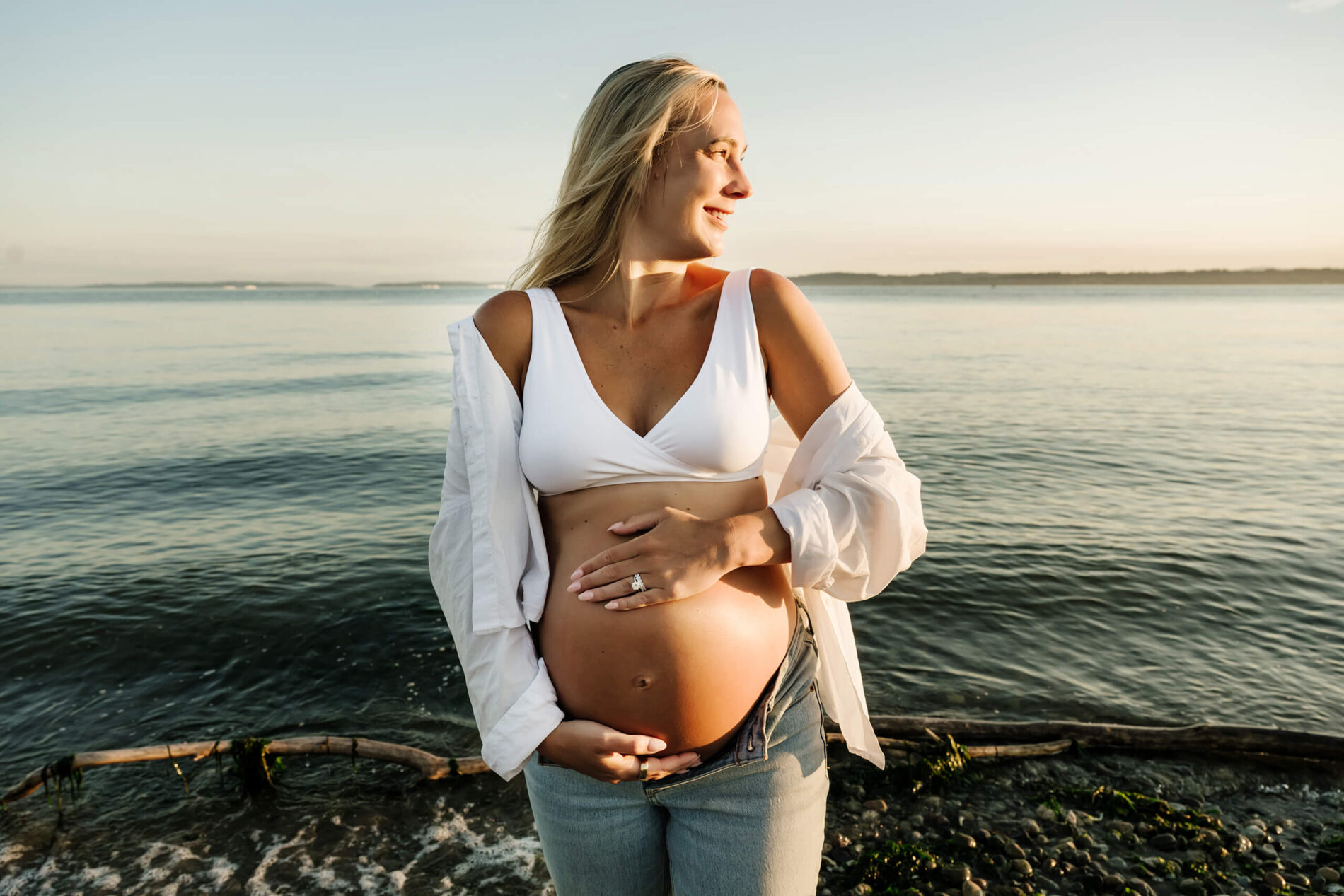 Pregnant woman standing by the water with her hands on her belly during an outdoor maternity portrait.
