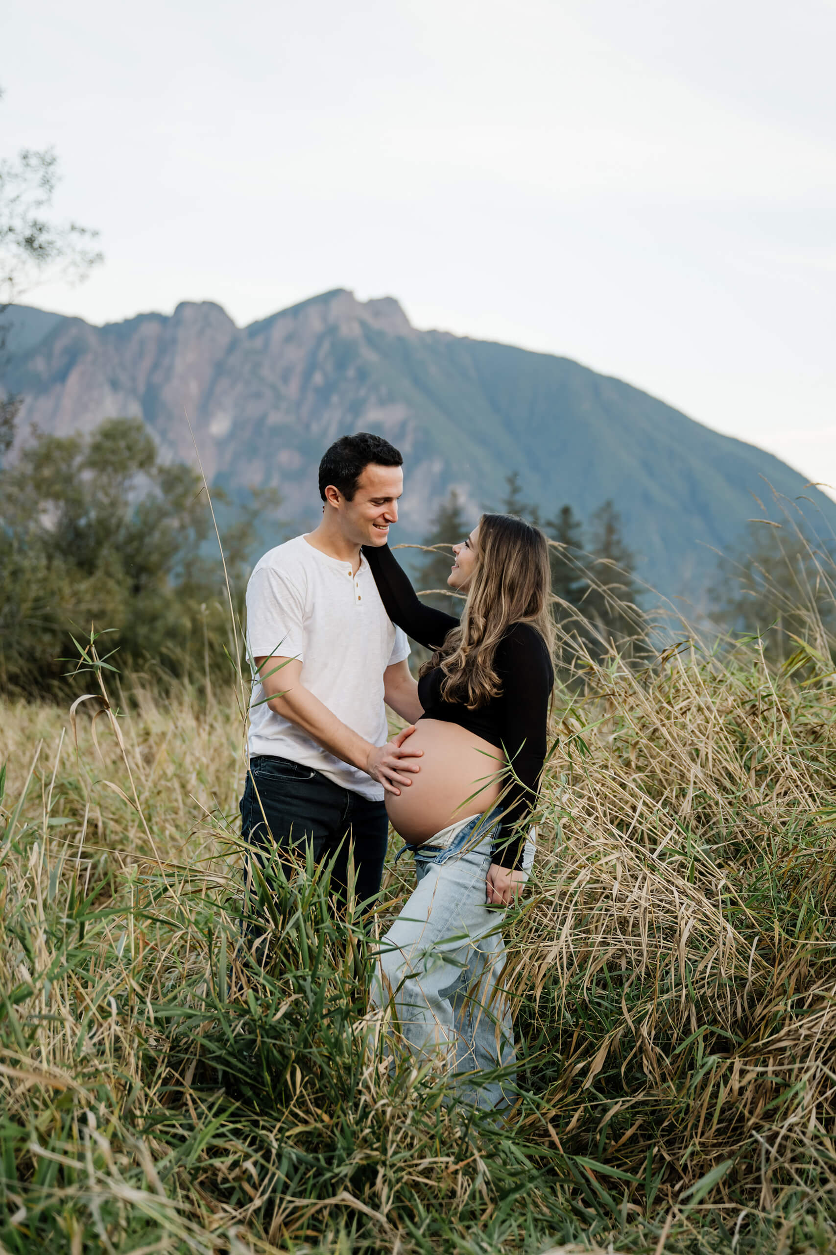 Expecting couple standing together in tall grass with mountains in the background.