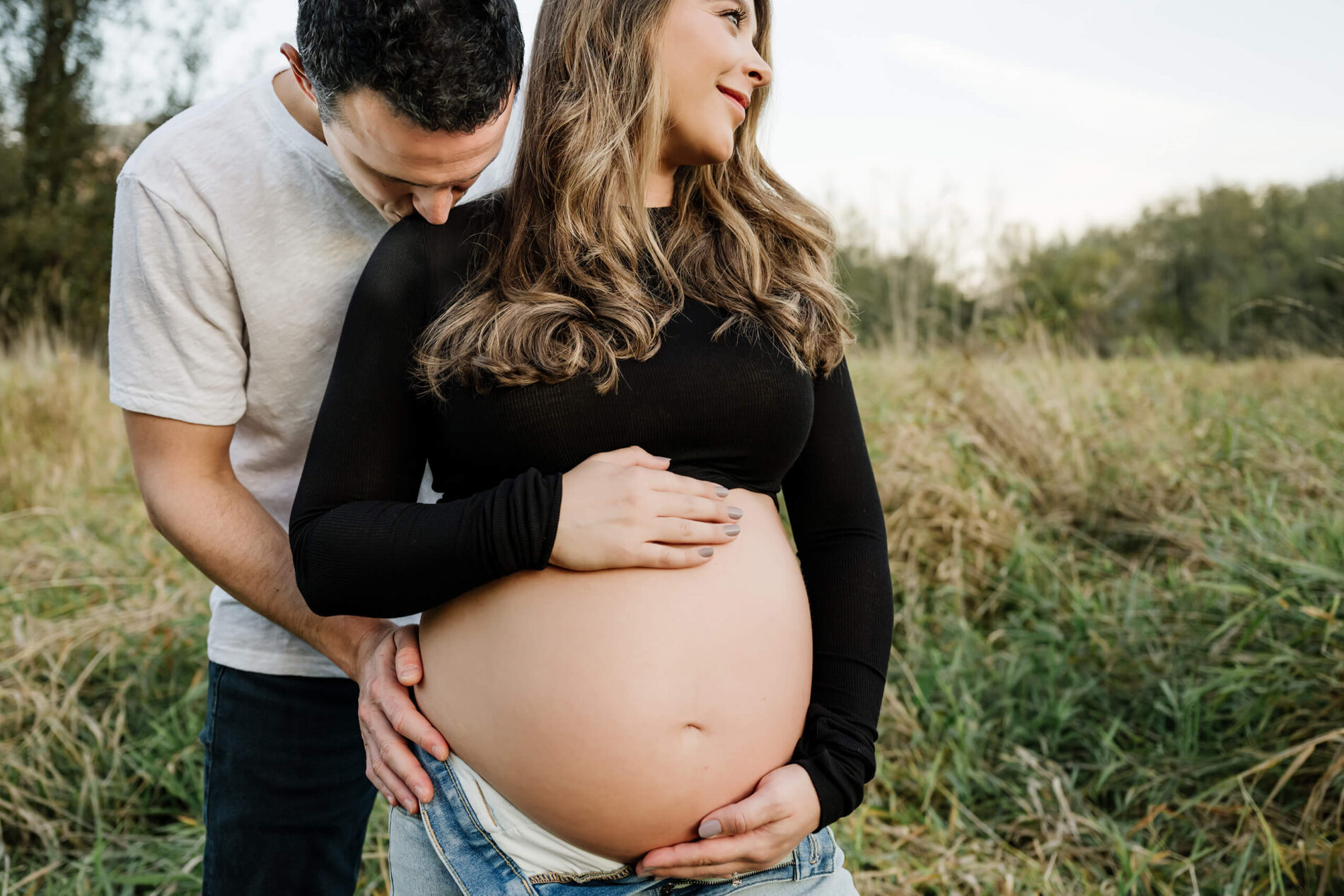 Expecting couple holding the baby bump in a grassy field during an outdoor maternity session.