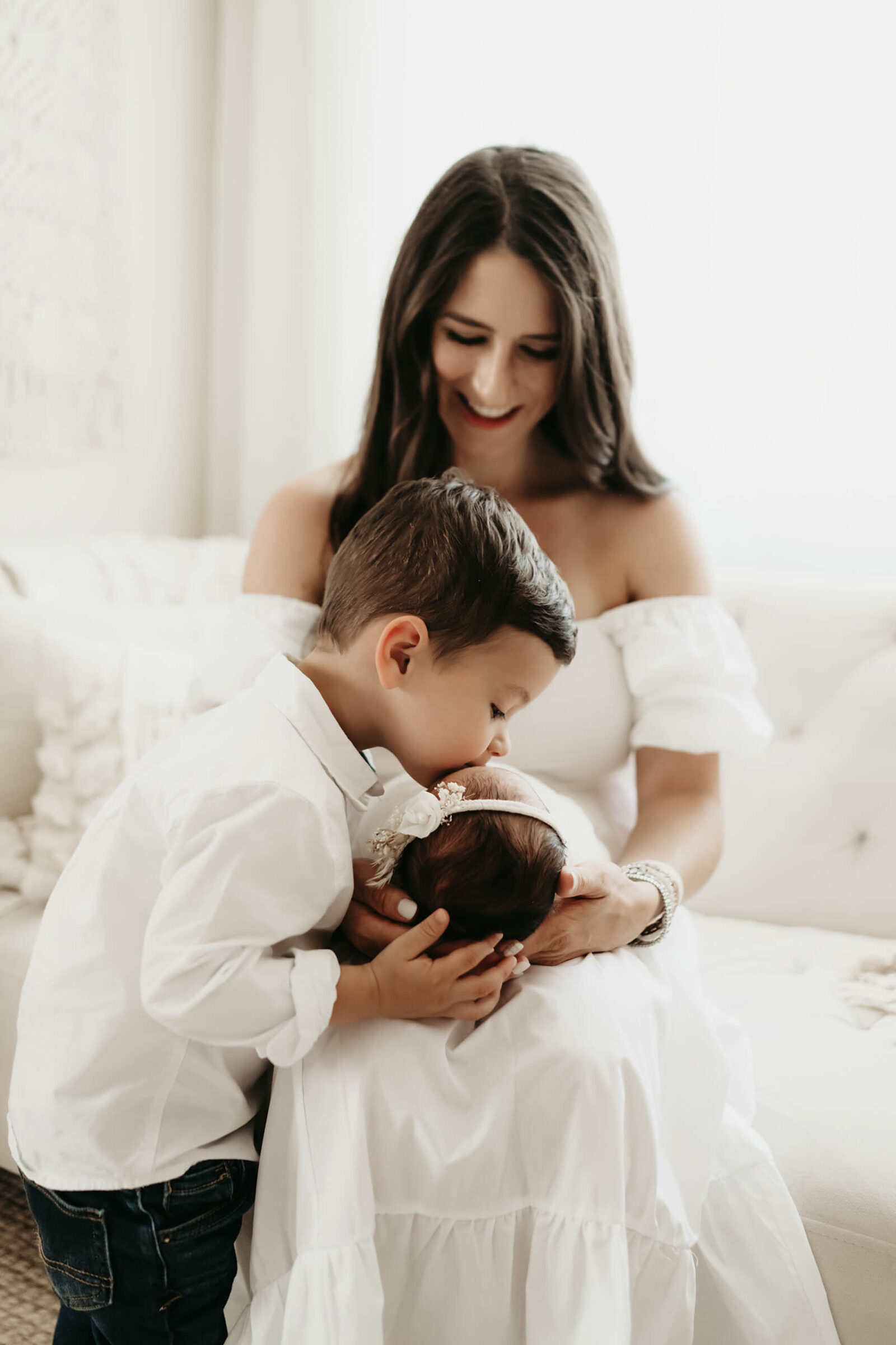 Big brother gently kissing his newborn baby sister while mom holds her during an in-home newborn session.