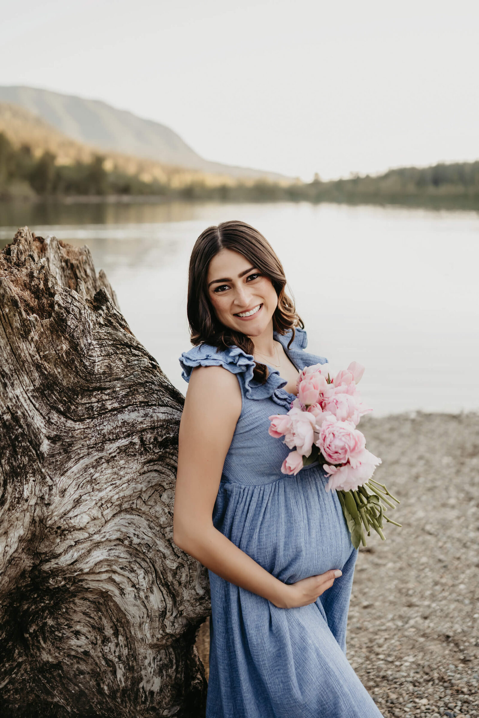 Expecting mom in a blue dress holding pink flowers by a mountain lake during a natural outdoor maternity session.