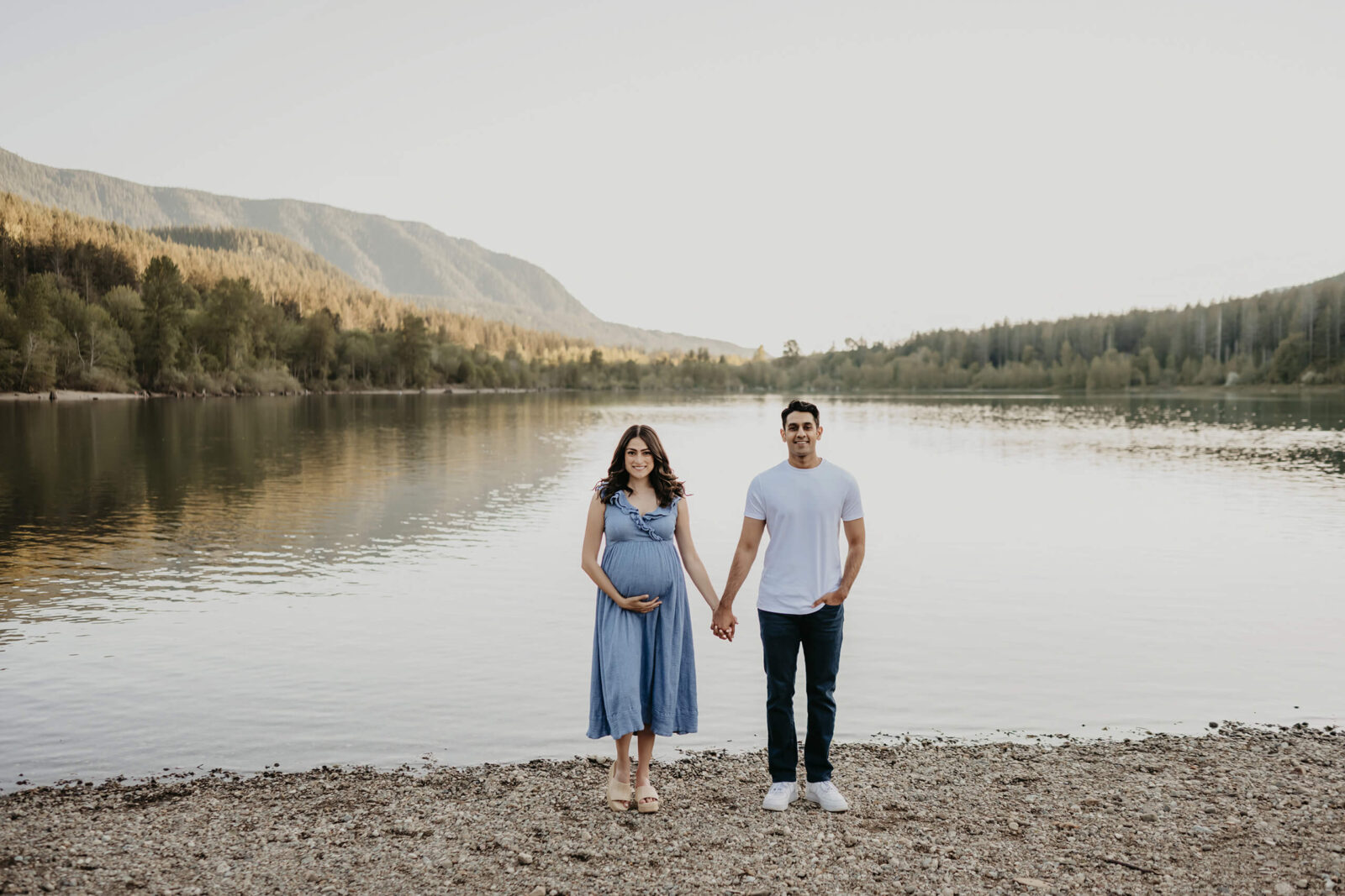 Expecting parents holding hands by a quiet mountain lake during a natural outdoor maternity session.