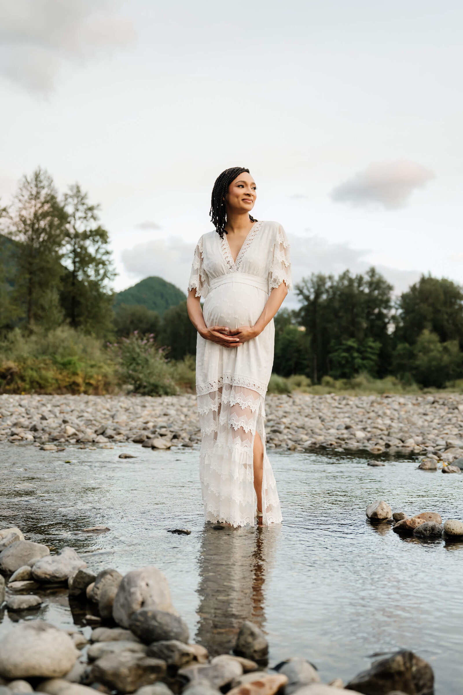 Expecting mother standing in shallow water with trees and mountains behind her during an outdoor maternity session.
